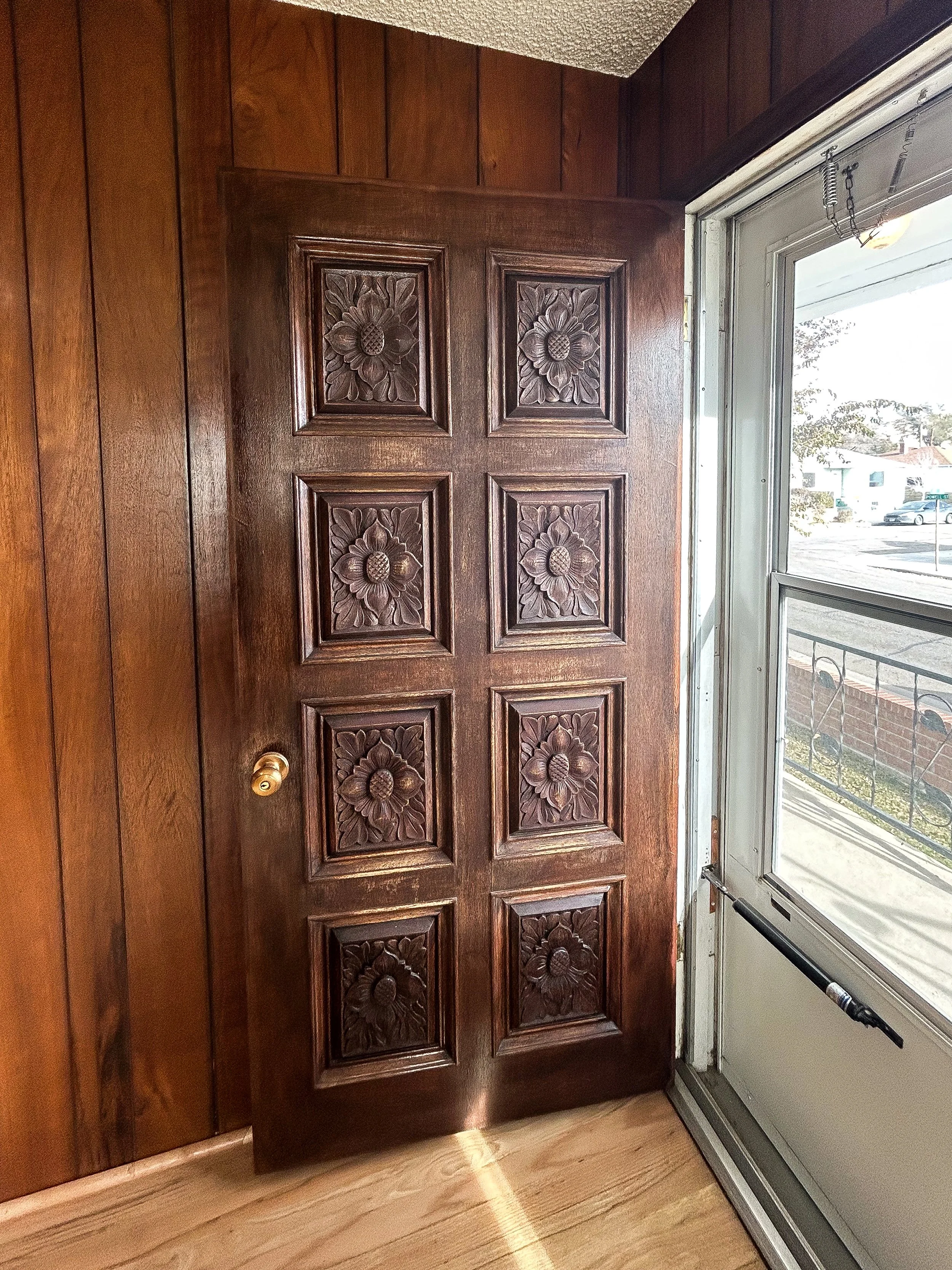 Wooden door with carved floral patterns next to a window in a room with wood-paneled walls.