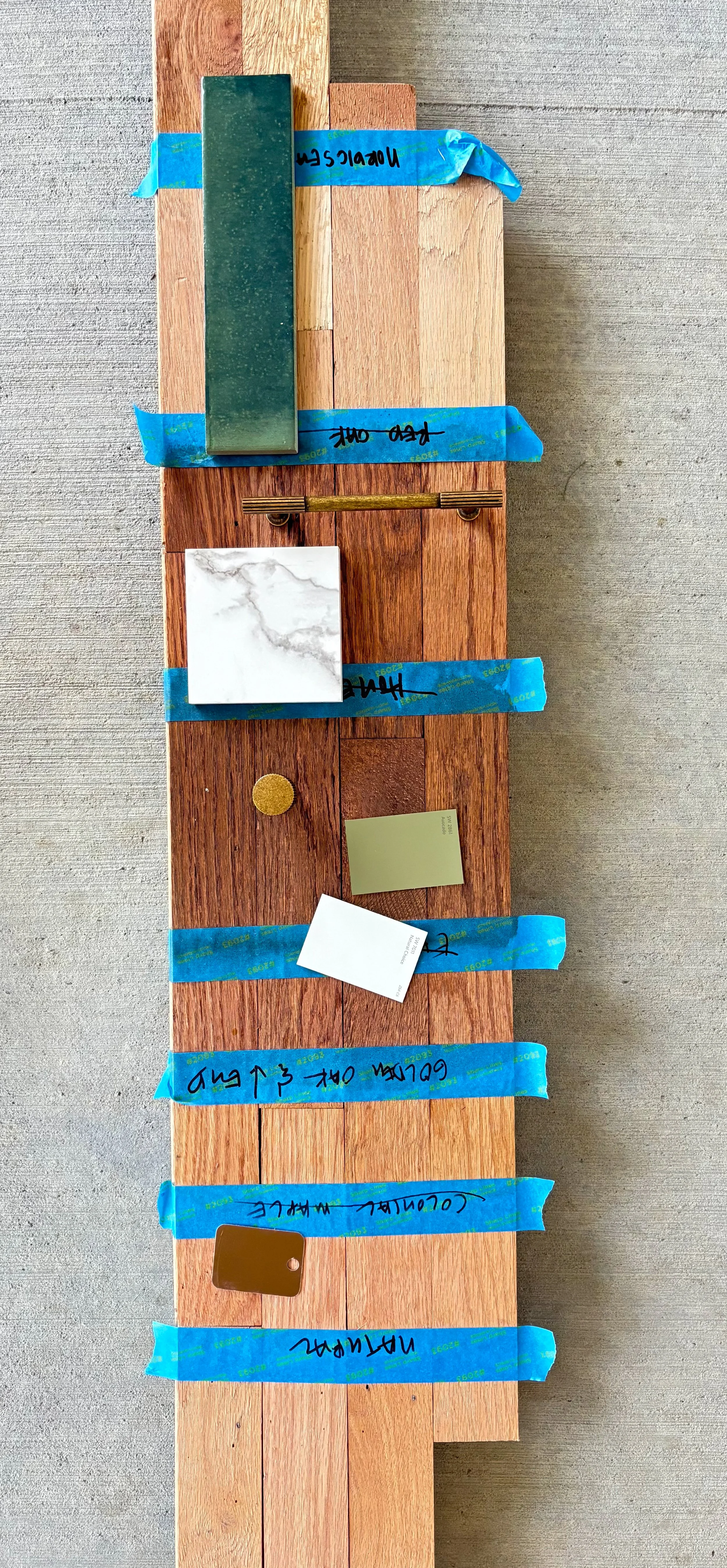 Swatch sample board with stones, tiles, and paint color swatches secured with blue painter's tape on a wooden surface.