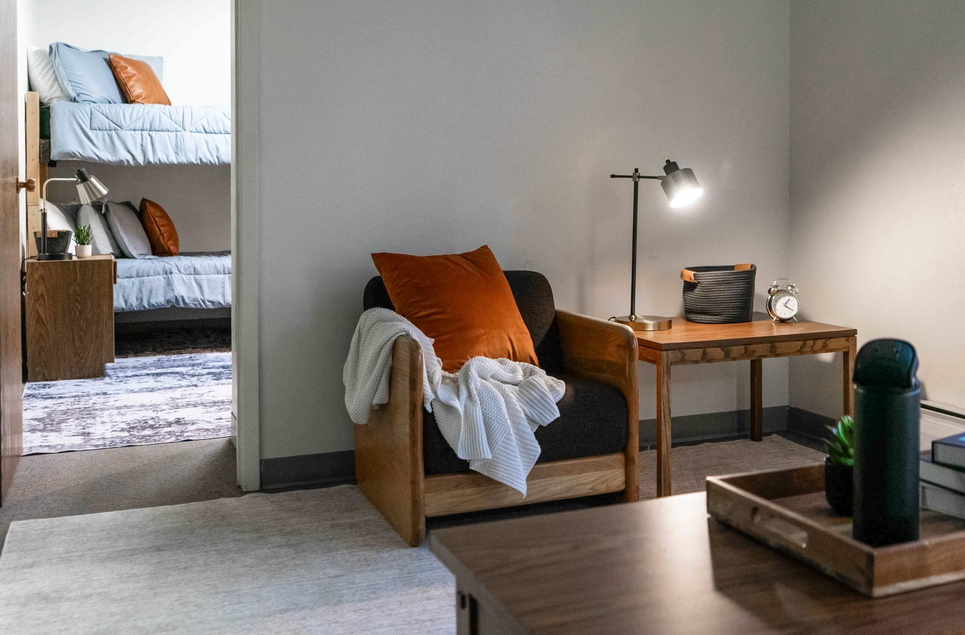 Living room with a black armchair that has an orange pillow and a white blanket, a wooden side table with a desk lamp, a basket, and an alarm clock, and in the background, there is a bedroom with bunk beds with blue bedding and orange pillows.