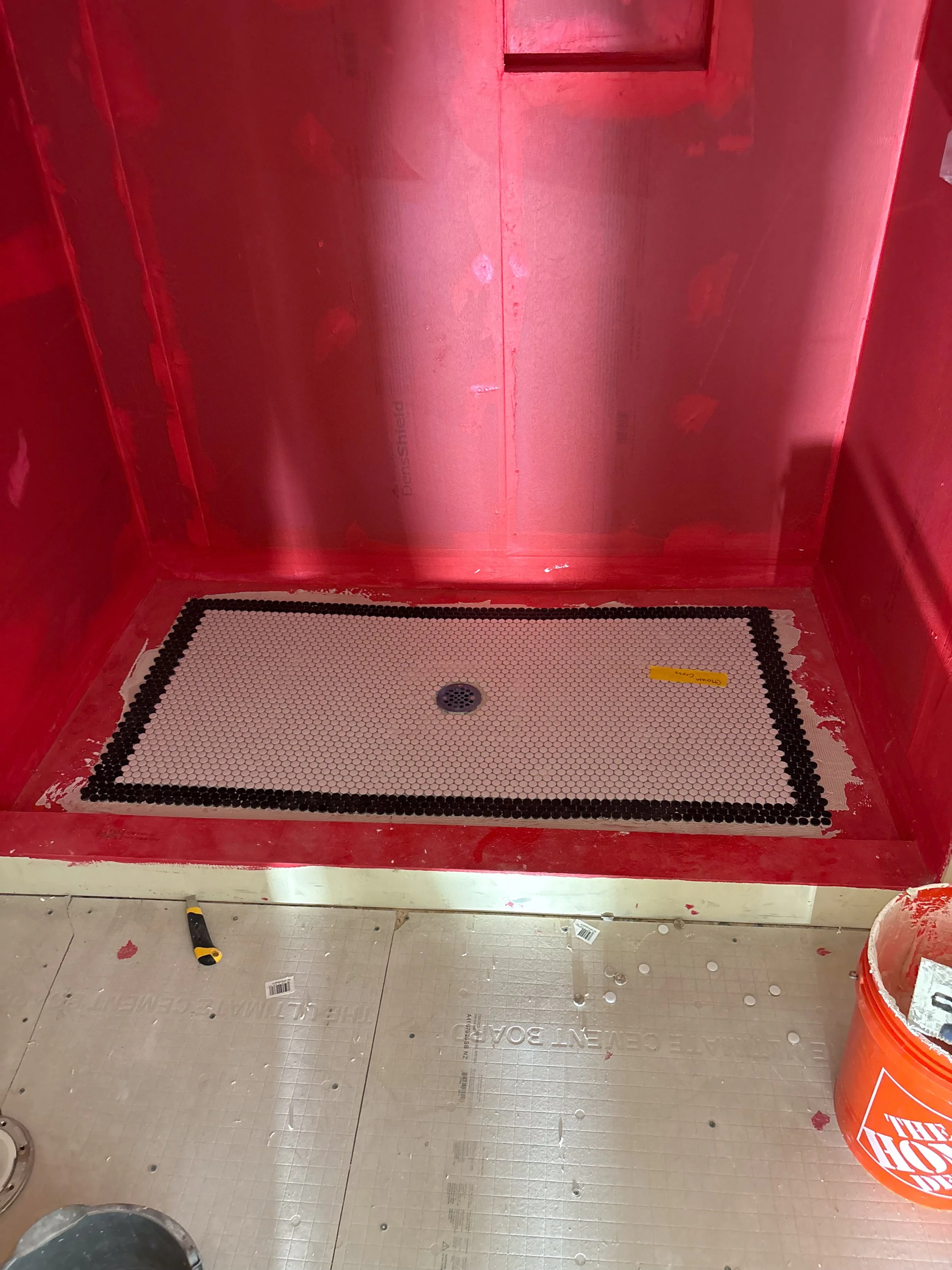 A shower stall with white penny tile outlined by black penny tile with a drain in the center, and a yellow utility knife on the floor.
