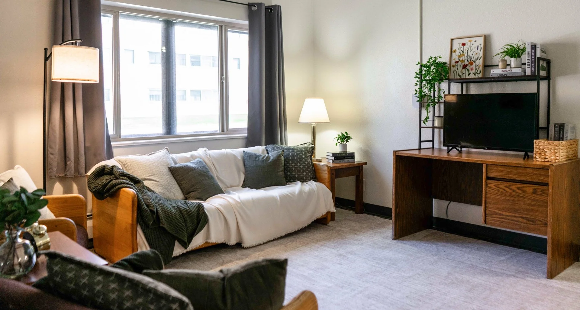 Living room with a sofa covered with a white throw, grey cushions, a side table with a lamp, a window with curtains, a wooden desk with a TV and shelving with decorative items and plants.