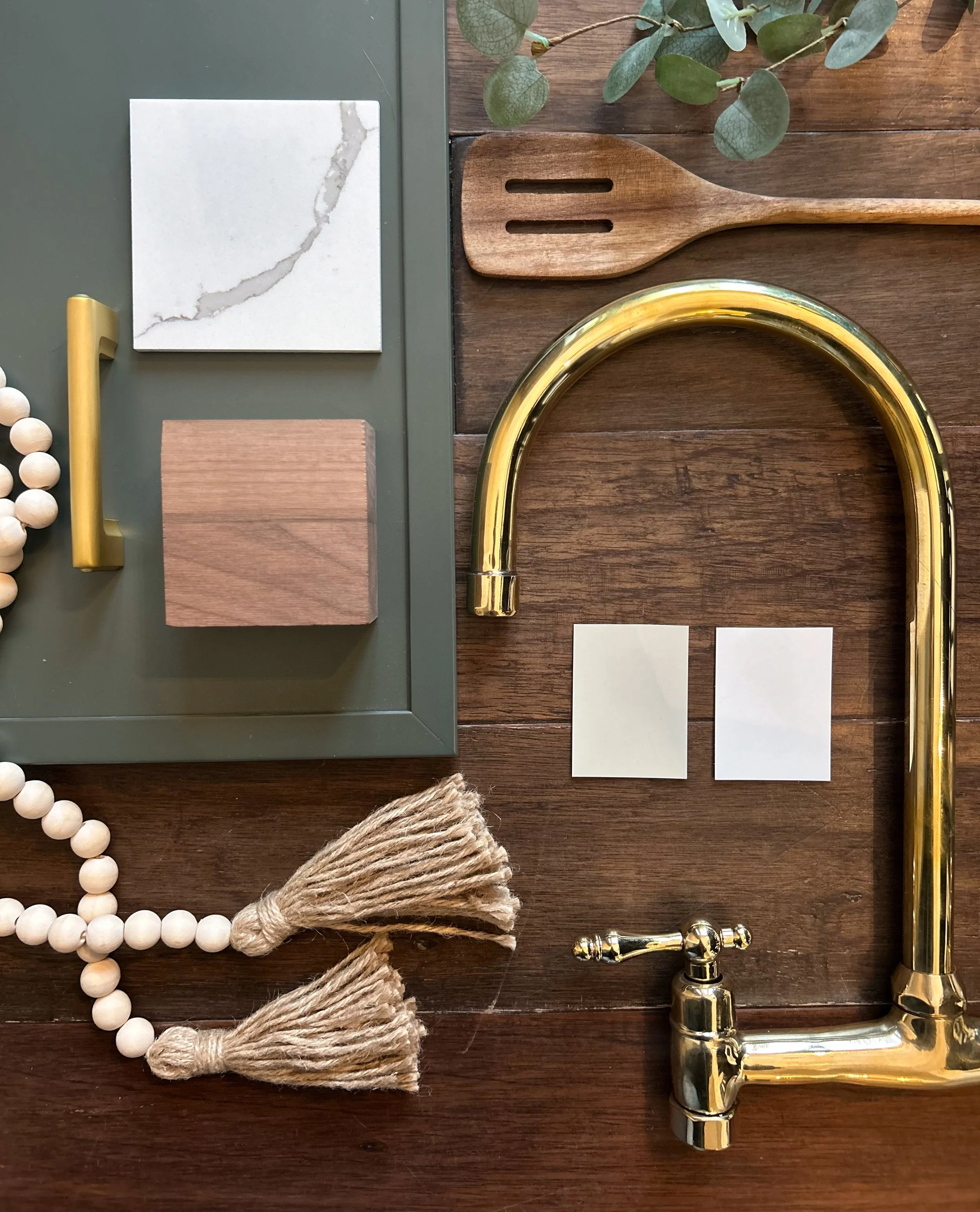 A flat lay of a kitchen or bathroom faucet with various sample materials and color swatches, greenery in the top right corner, and decorative beads with tassels on a wooden surface.