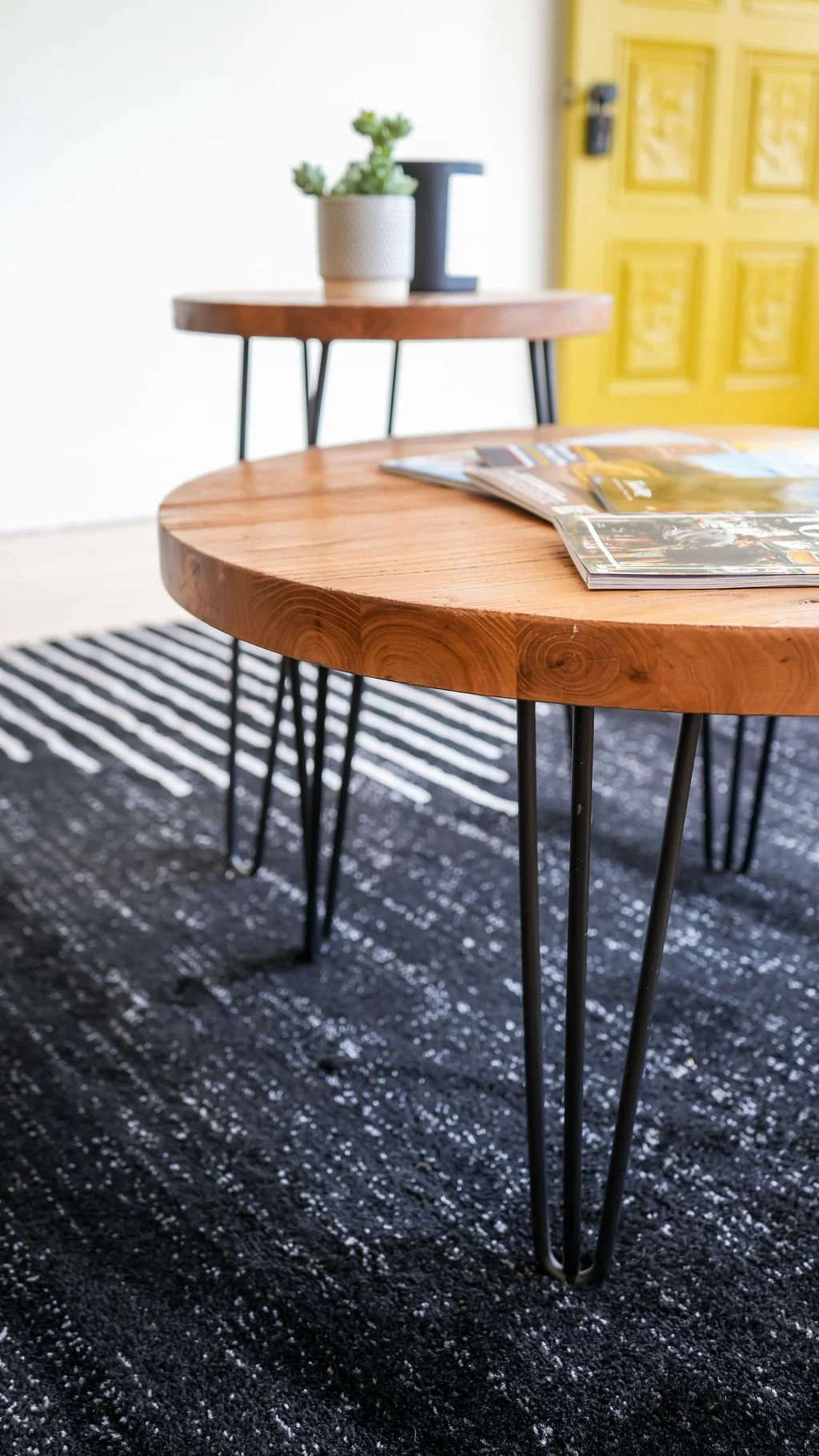Close-up of a round wooden table with black metal hairpin legs, placed on a black and white patterned rug, with magazines on top. In the background, there's a smaller wooden table with a potted succulent plant and a black decorative object, against a yellow cabinet door.