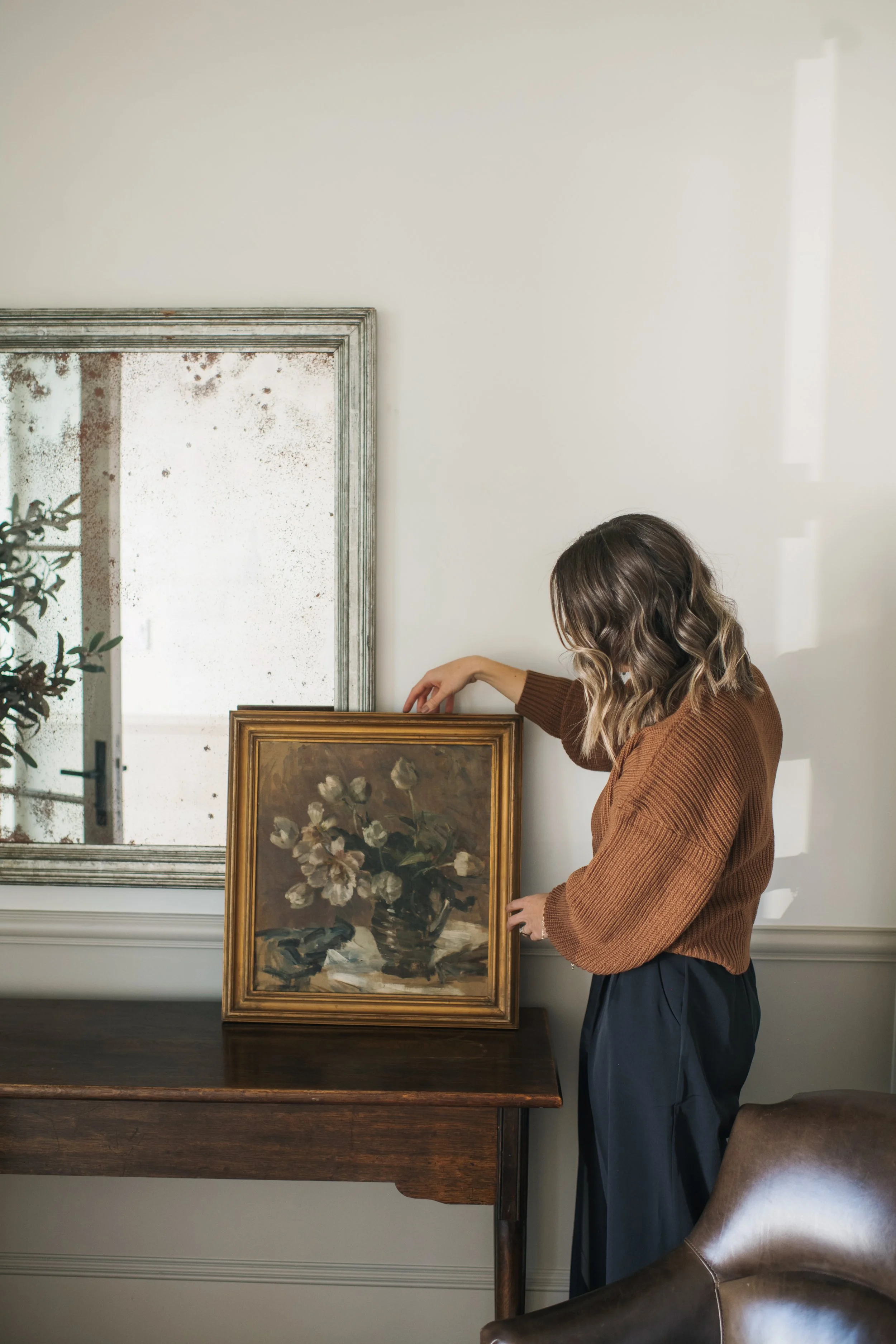 A woman with wavy hair, wearing a brown sweater and dark pants, is standing in a room, adjusting a framed oil painting of white flowers in a vase on a wooden table.