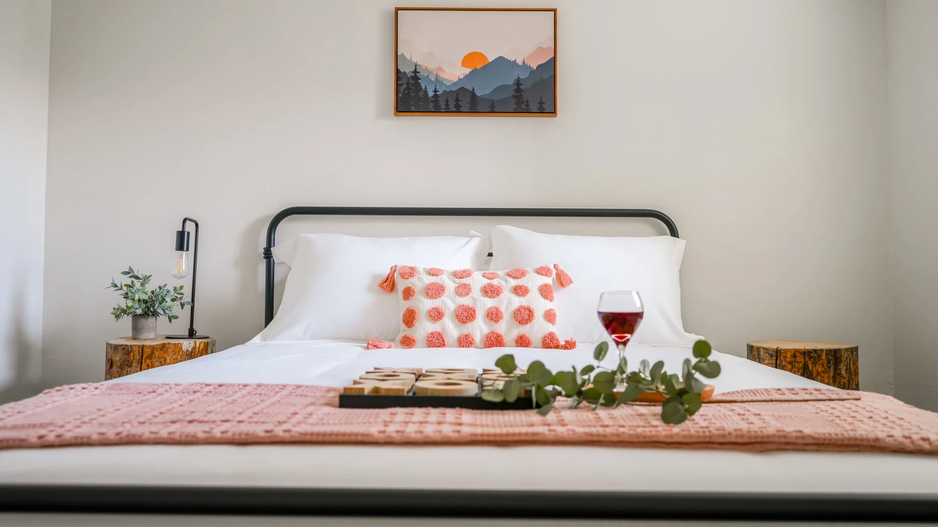 A neatly made bed with white sheets and a pink textured blanket, adorned with a decorative pillow with pink pom-poms, set against a plain white wall with a mountain landscape painting above. There are two matching wooden bedside tables with a lamp and plant, and a glass of red wine and a tray with word blocks and eucalyptus on the bed.