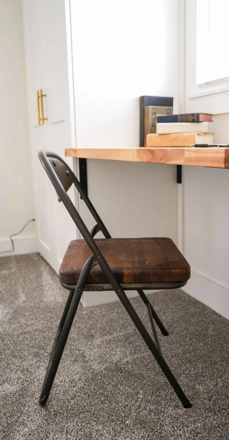 A black iron folding chair with a wooden seat in front of a built-in wooden desk with books and a window in the background.