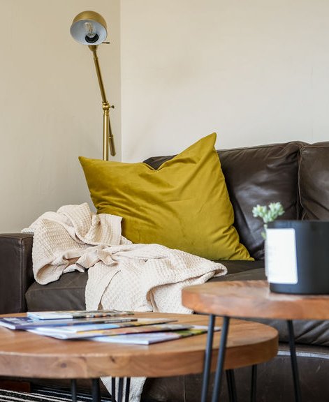 Living room with a brown leather sofa, a large yellow pillow, a cream blanket, a wooden coffee table with magazines, and a gold floor lamp.