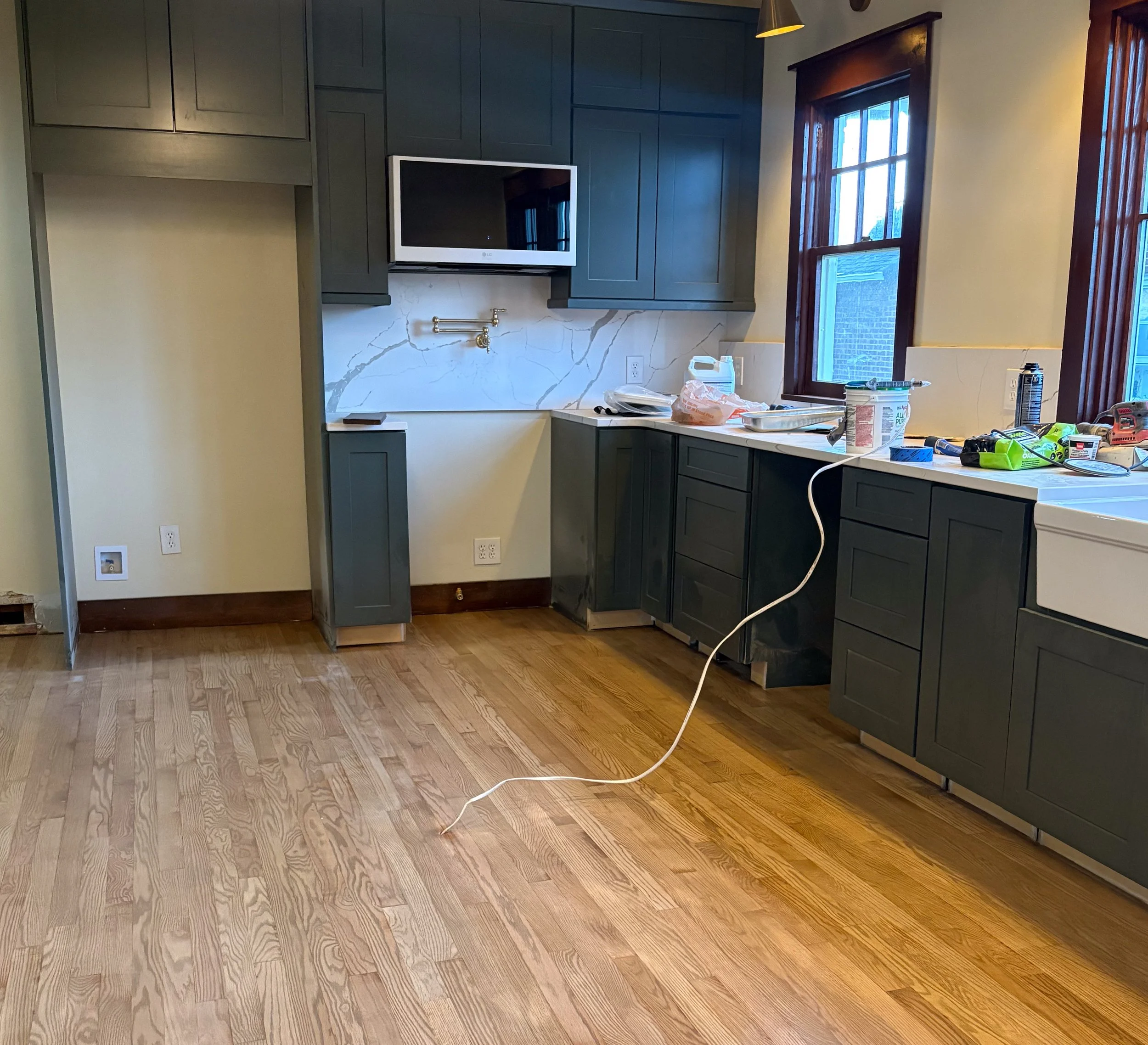 A kitchen with unfinished cabinetry and a wooden floor, cluttered with tools and supplies, and featuring two large windows with wooden frames.