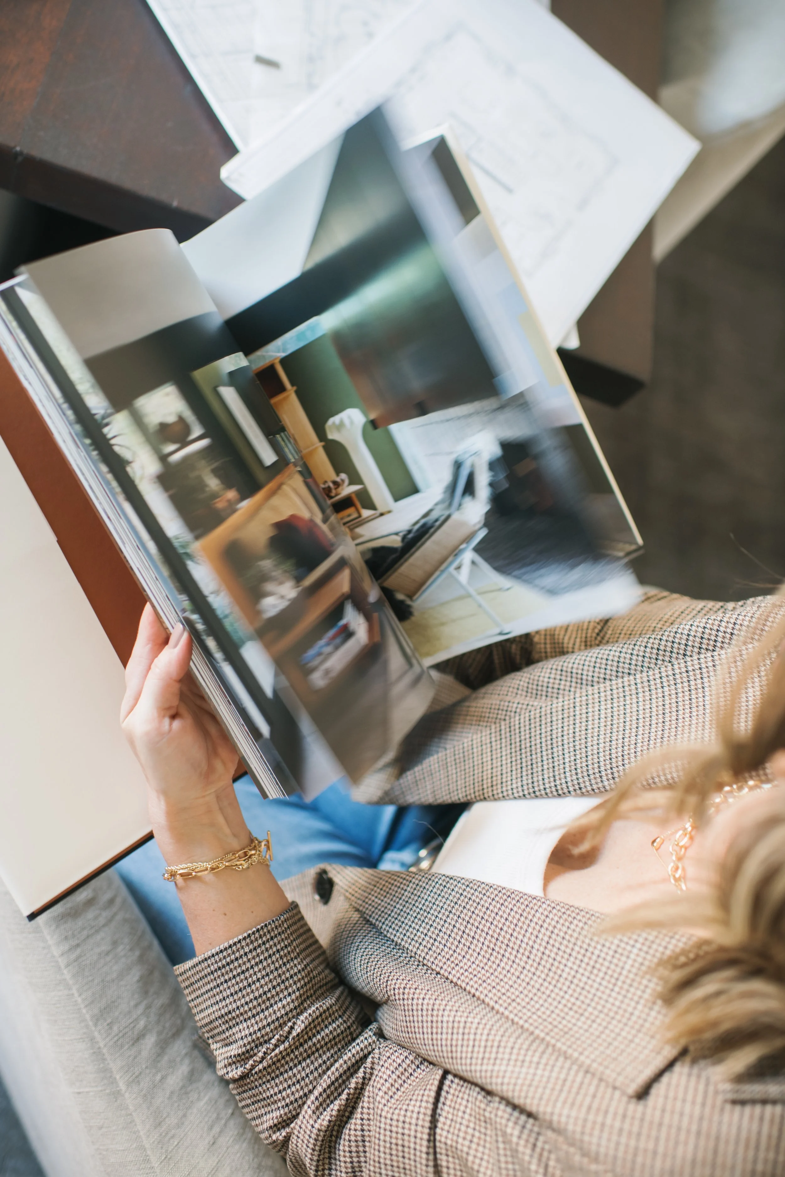 Person holding an open magazine with images of interior design and decor, sitting on a beige chair at a dark wooden table.