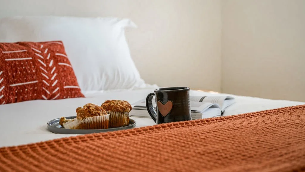 A neatly made bed with white sheets, a white pillow, and a decorative burnt orange pillow with white patterns. On the bed, there is a tray with two muffins, an open book, and a black mug with a heart design.