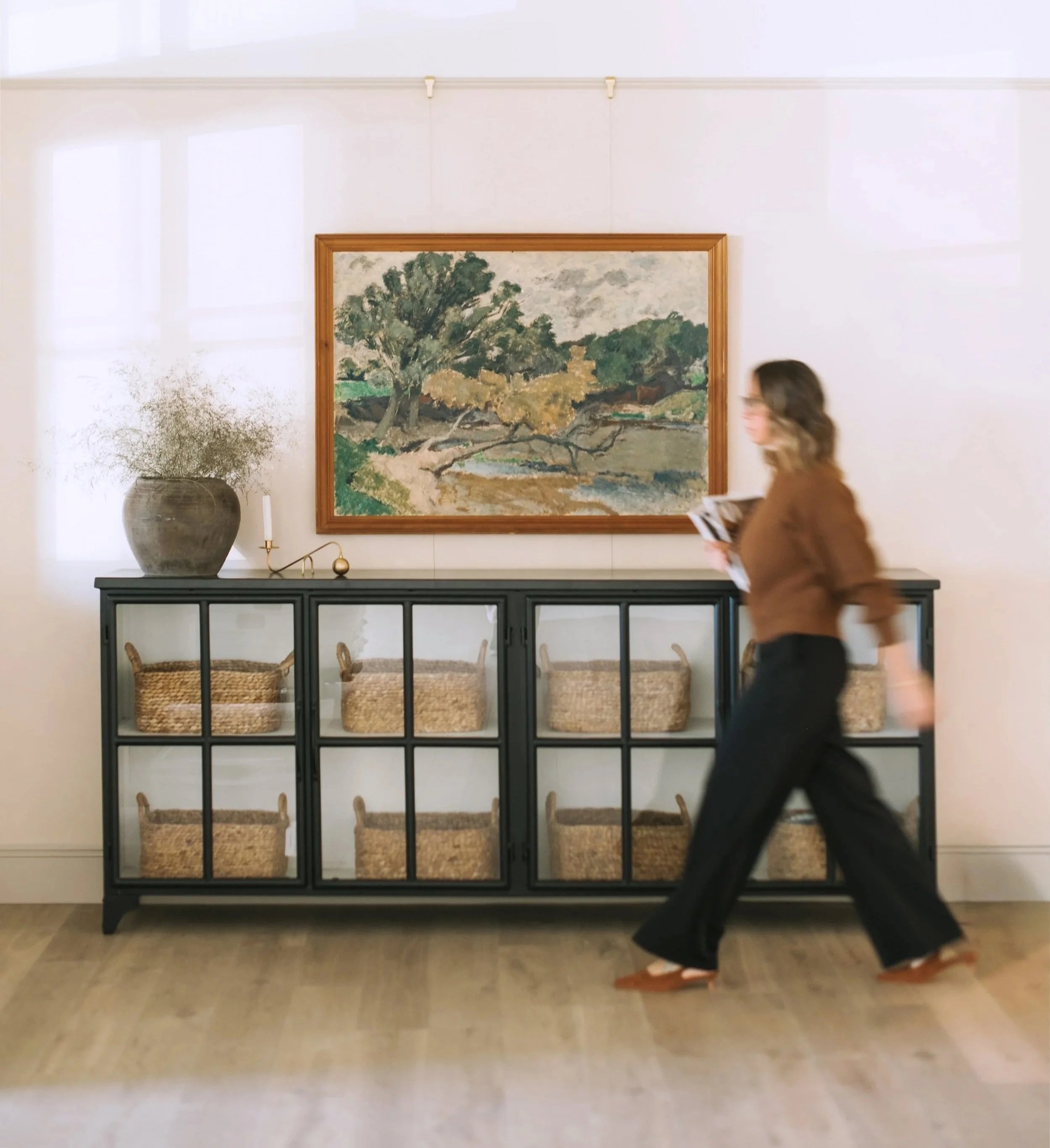 A woman walking past a black console table with six wicker baskets, a large vase with dried plant, and a small candle holder, in front of a white wall with a landscape painting.
