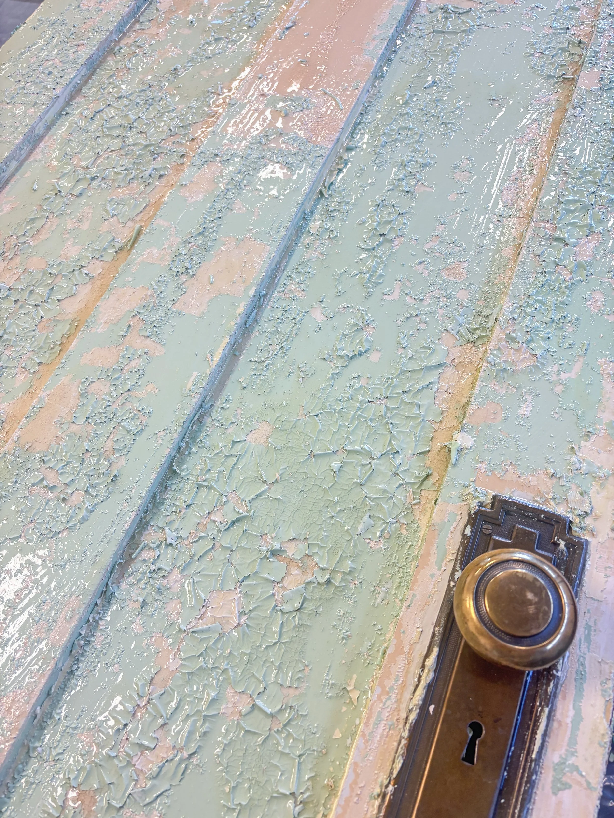 Close-up of a wooden door with peeling pale green paint and a brass doorknob.