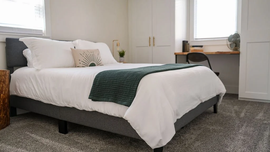 A neatly made bed with white bedding, grey headboard, green throw blanket, decorative pillow, and a small potted plant on a bedside table in a bright bedroom.