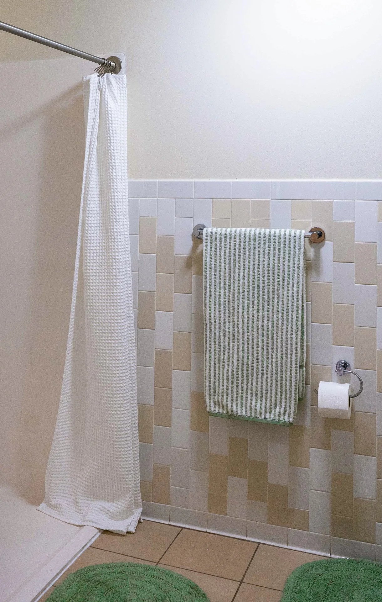 Bathroom with beige and white tiles, towel rails with a green-striped towel, a toilet paper roll, and green rugs on a tiled floor.