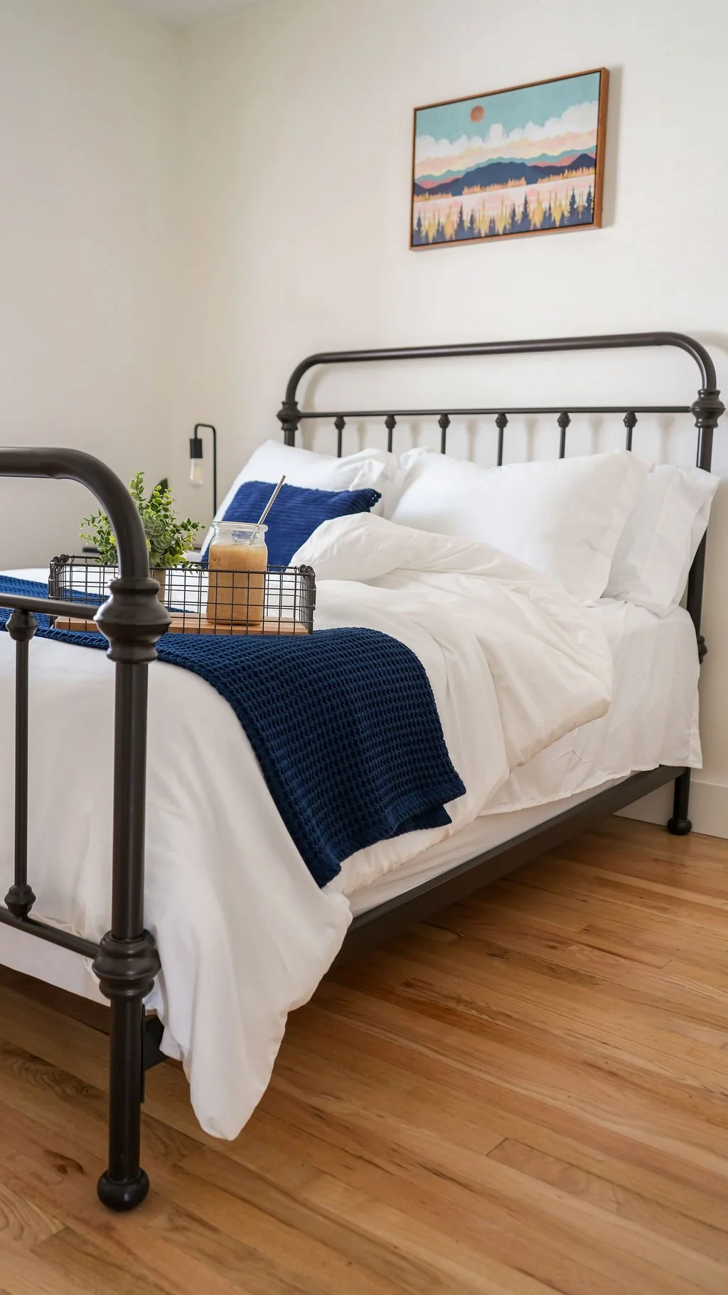 A neatly made bed with white bedding, a navy blue throw blanket, and matching navy blue pillow in a bedroom with wooden flooring and a colorful landscape painting on the wall.