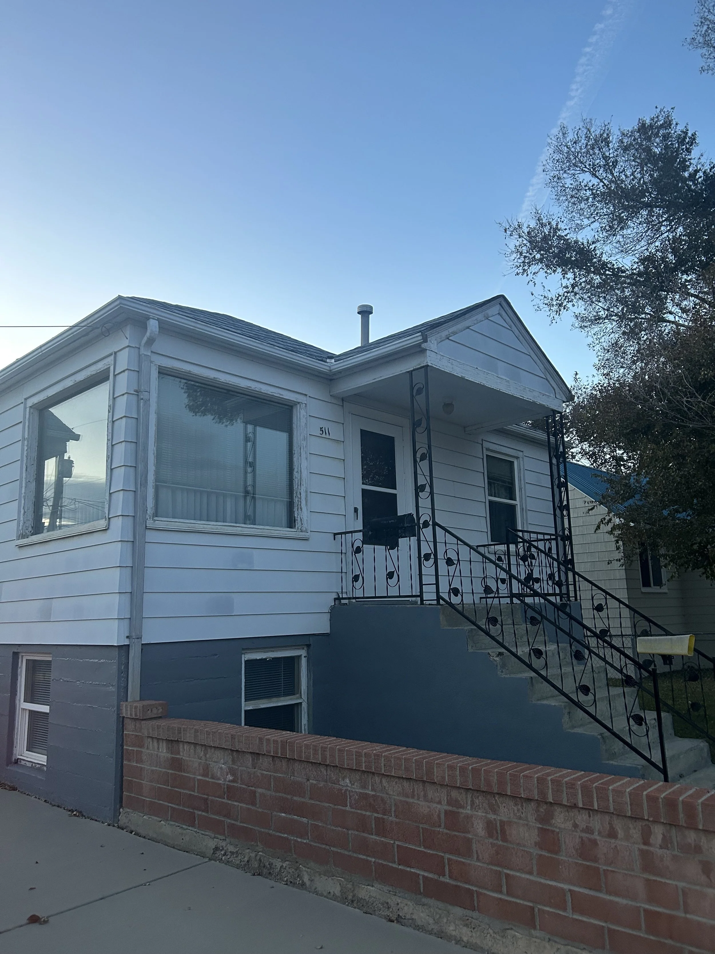 Front view of a small, light blue house with a porch and black wrought iron railing, stairs leading up to a door, a large window with white trim, and a brick wall at the sidewalk. There are trees and a clear blue sky in the background.