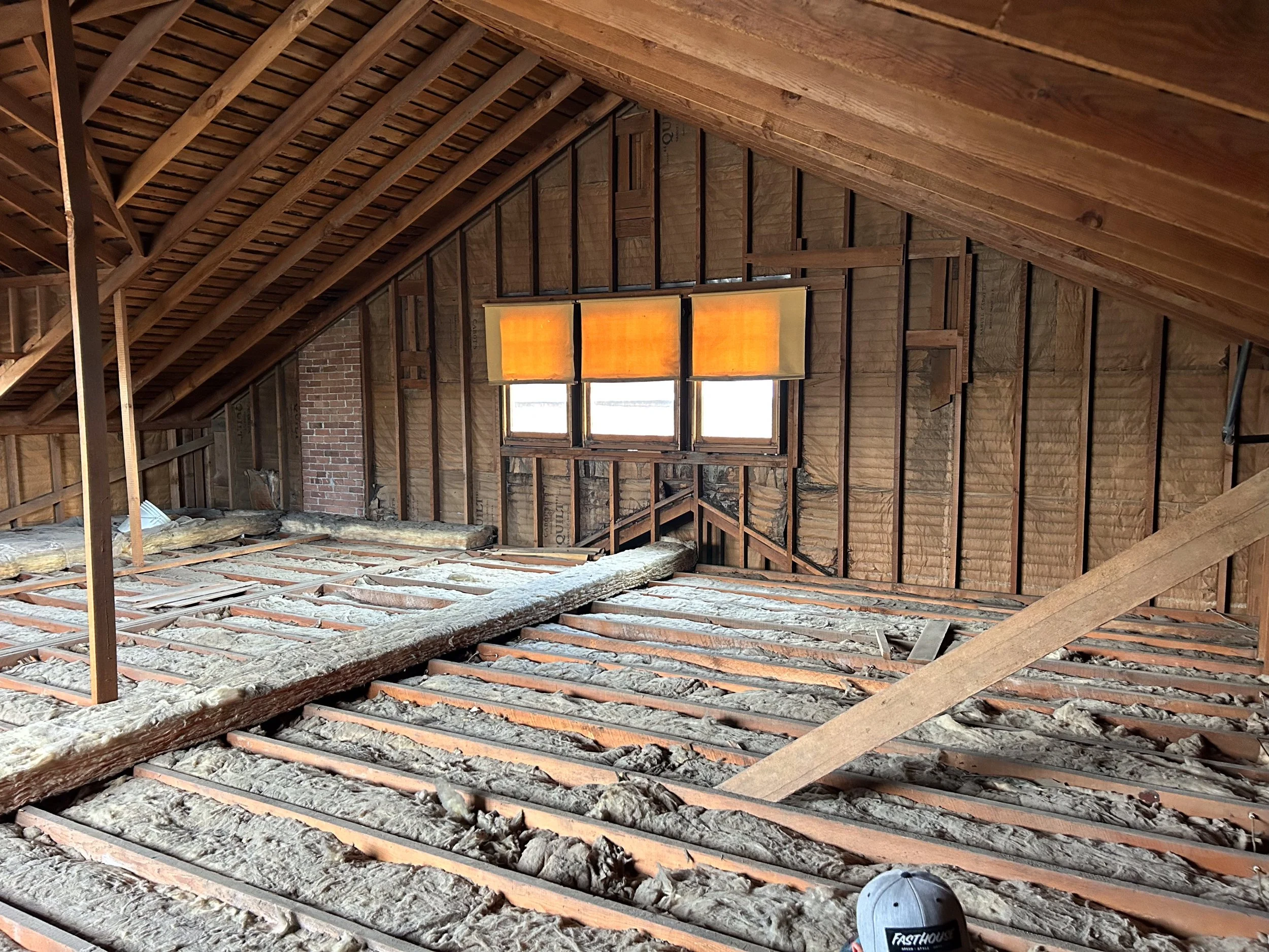 An attic in the process of renovation, showing exposed wooden beams and insulation on the floor, with a window covered by a brown shade.
