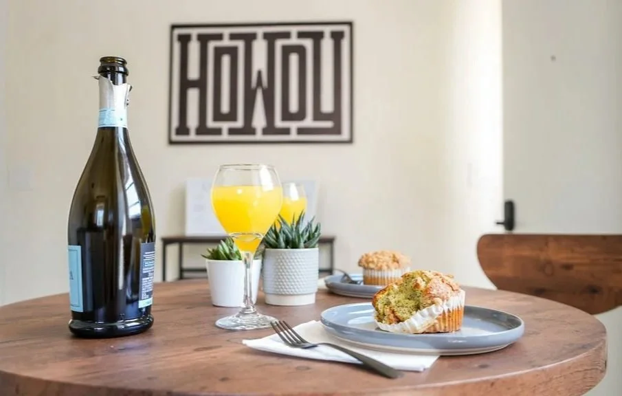 A table with a bottle of champagne, two glasses of orange juice, two cupcakes, a fork, and a plate with a slice of cake in front of a wall art that says 'Howdy'.