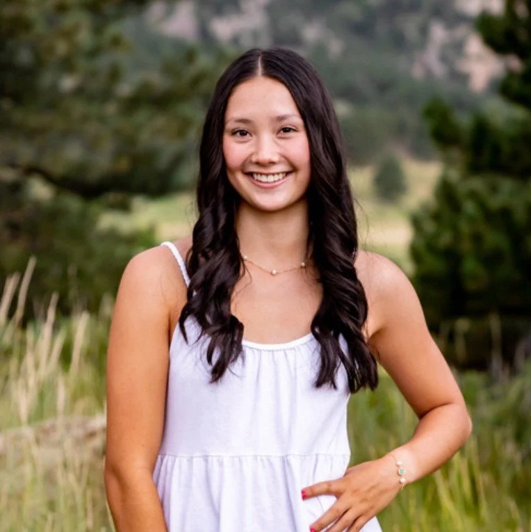 A young woman with long dark wavy hair wearing a white sleeveless dress, standing outdoors in a grassy area with trees in the background, smiling at the camera.