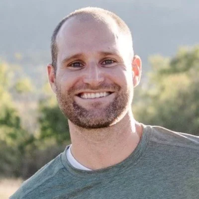 A smiling man with a short beard and buzz cut hairstyle outdoors, with trees and a mountain in the background.