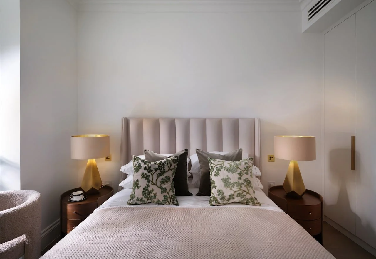 A neatly made bed with an upholstered headboard, two side tables with beige lamps, decorative cushions, and a cup on a tray, in a modern bedroom with minimal decor.