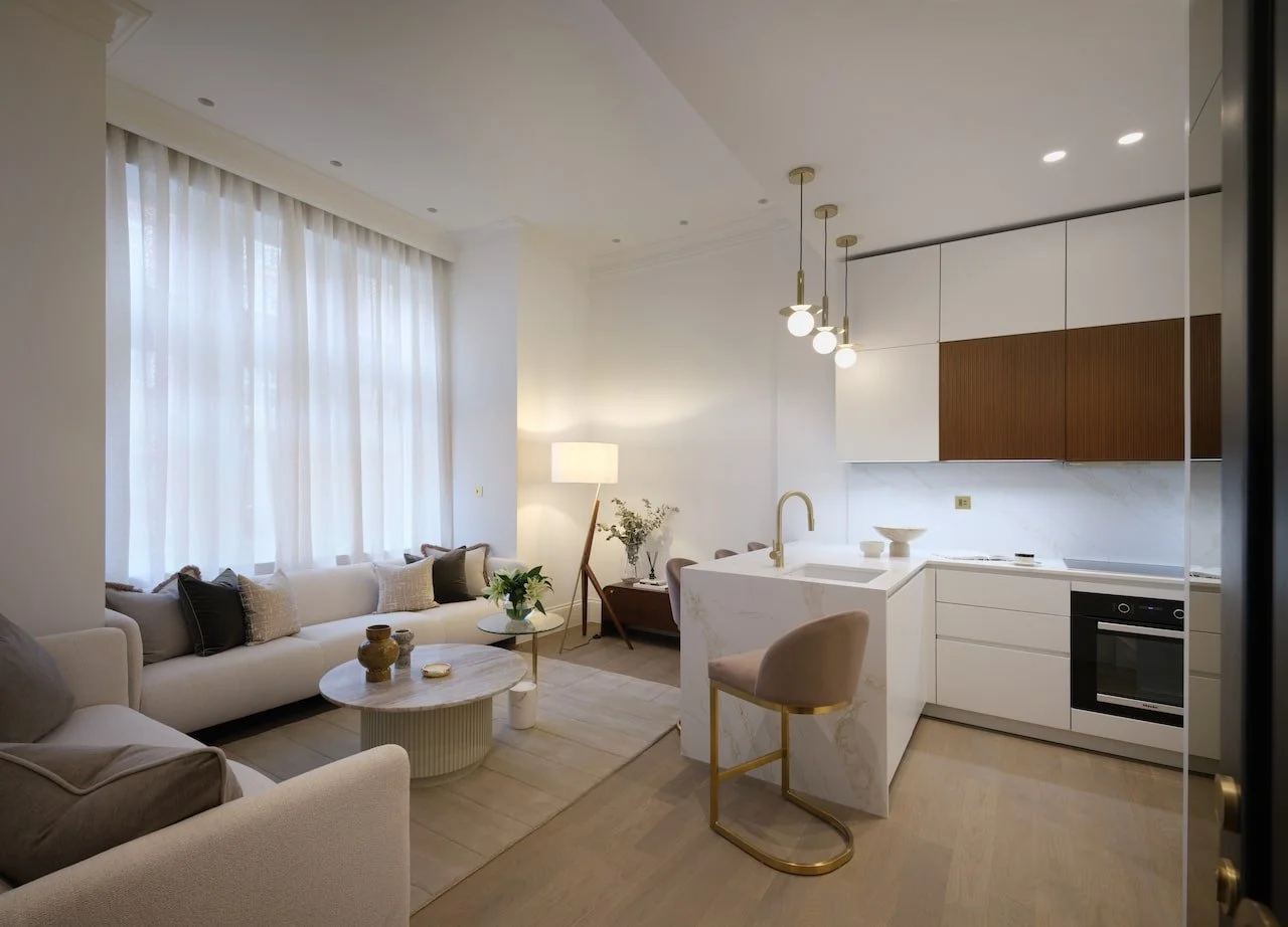 Open-concept living room and kitchen with modern decor, beige sofas, wooden and white cabinetry, marble countertops, and pendant lighting.