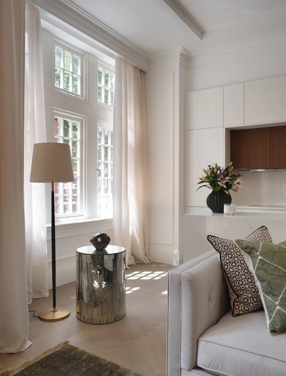 Living room with large windows, white walls, beige curtains, a beige sofa with patterned pillows, a tall floor lamp, a round mirrored side table with a small decorative object, and a black vase with flower arrangement on the built-in cabinet.