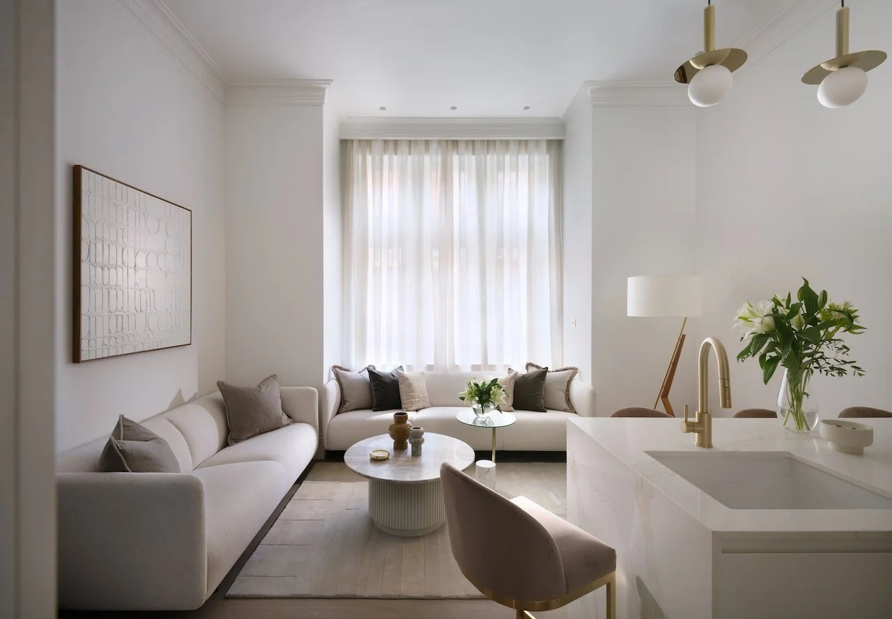 Bright living room with white sofas, beige and black pillows, a round coffee table, large window with sheer curtains, modern floor lamp, minimalistic decor, and a small kitchen island with a vase of white flowers.