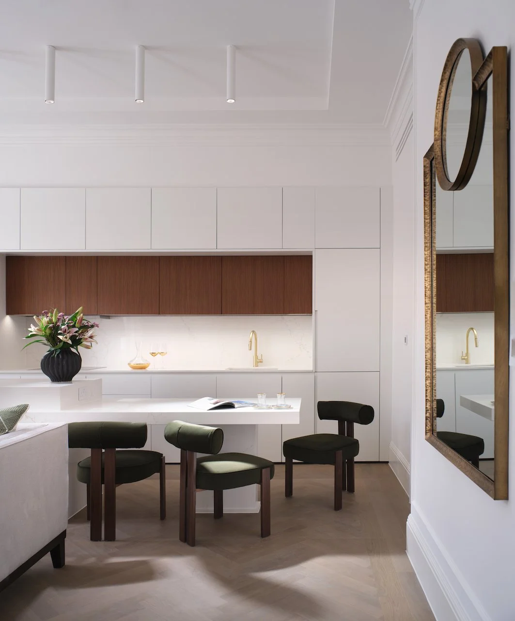 Modern kitchen with white cabinetry, a wooden backsplash, a gold faucet, black chairs, a white table with an open magazine and glasses, a vase with flowers, and a large mirror on the wall.