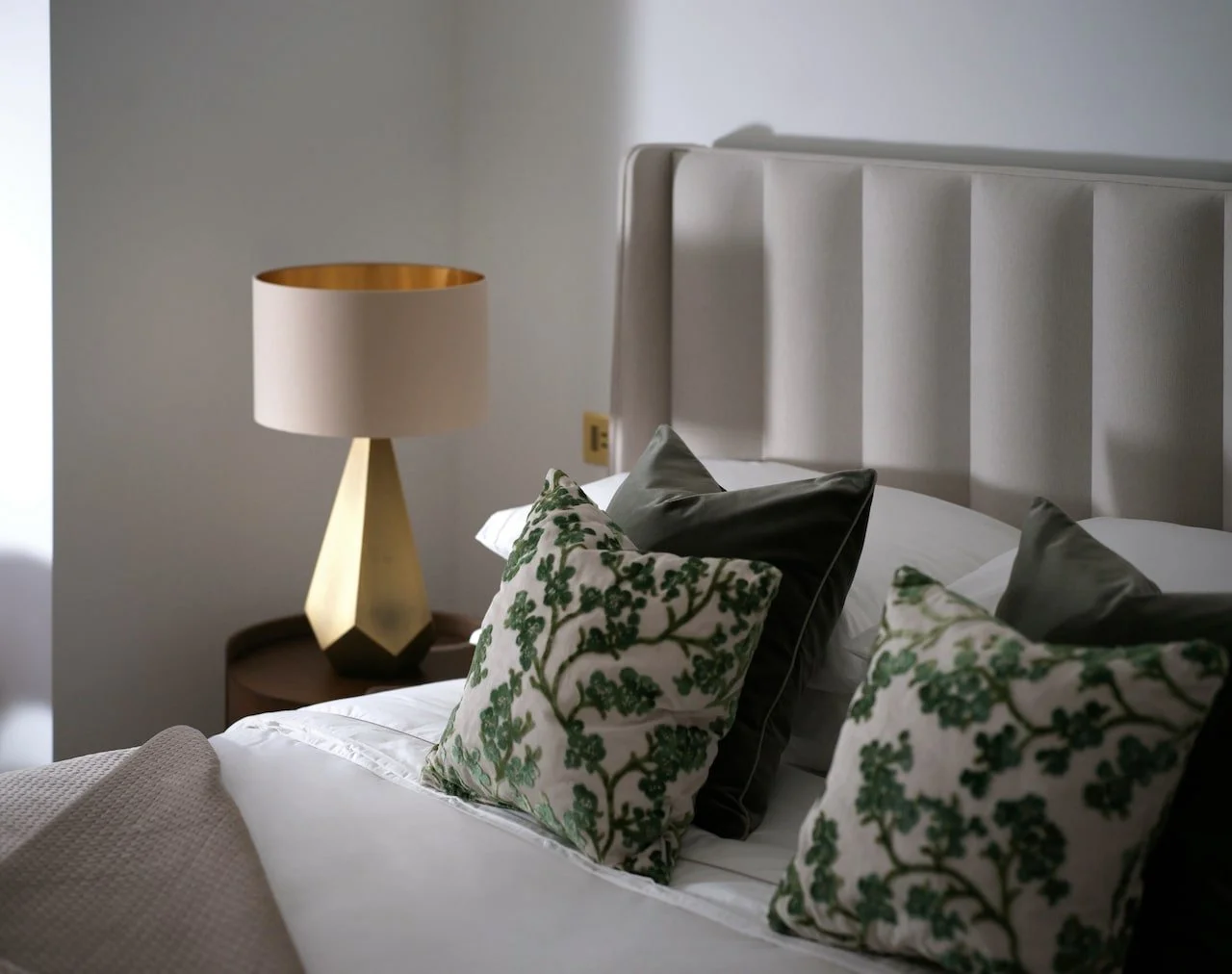 A neatly made bed with floral and dark green pillows, and a modern beige lamp on a wooden nightstand beside a white headboard in a minimalist bedroom.
