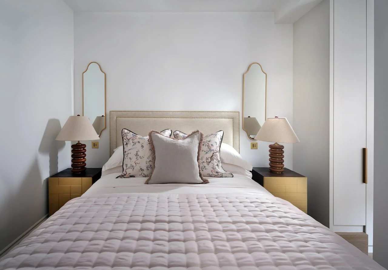 A neatly made bed with a beige headboard, two patterned pillows, and a smaller solid-colored pillow, flanked by two matching bedside tables with beige lamps, in a minimalist bedroom with white walls and built-in closet.