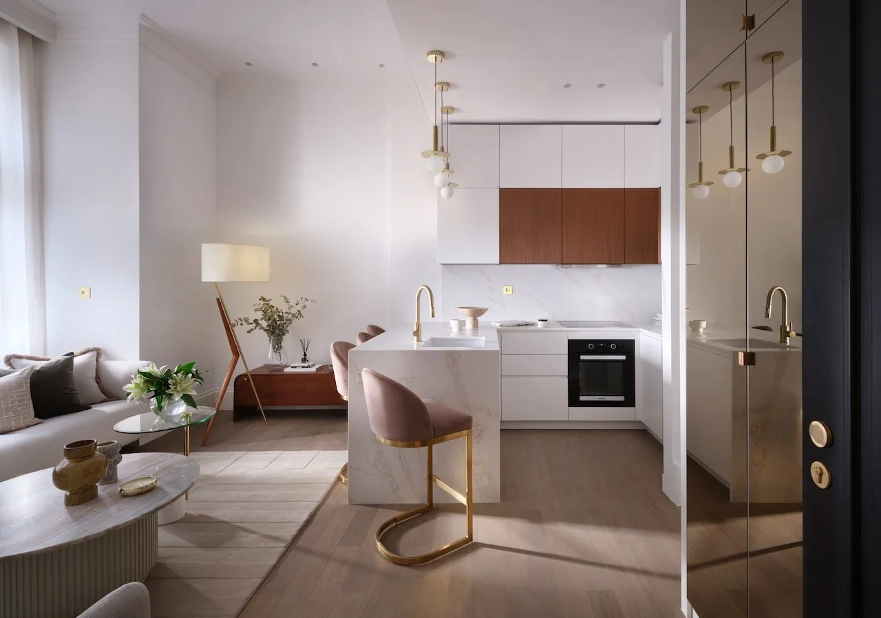 Modern open-concept living room and kitchen with white walls, wooden accents, and gold fixtures. The living room has a sofa, floor lamp, and coffee table, while the kitchen features a marble island, built-in oven, and hanging pendant lights.