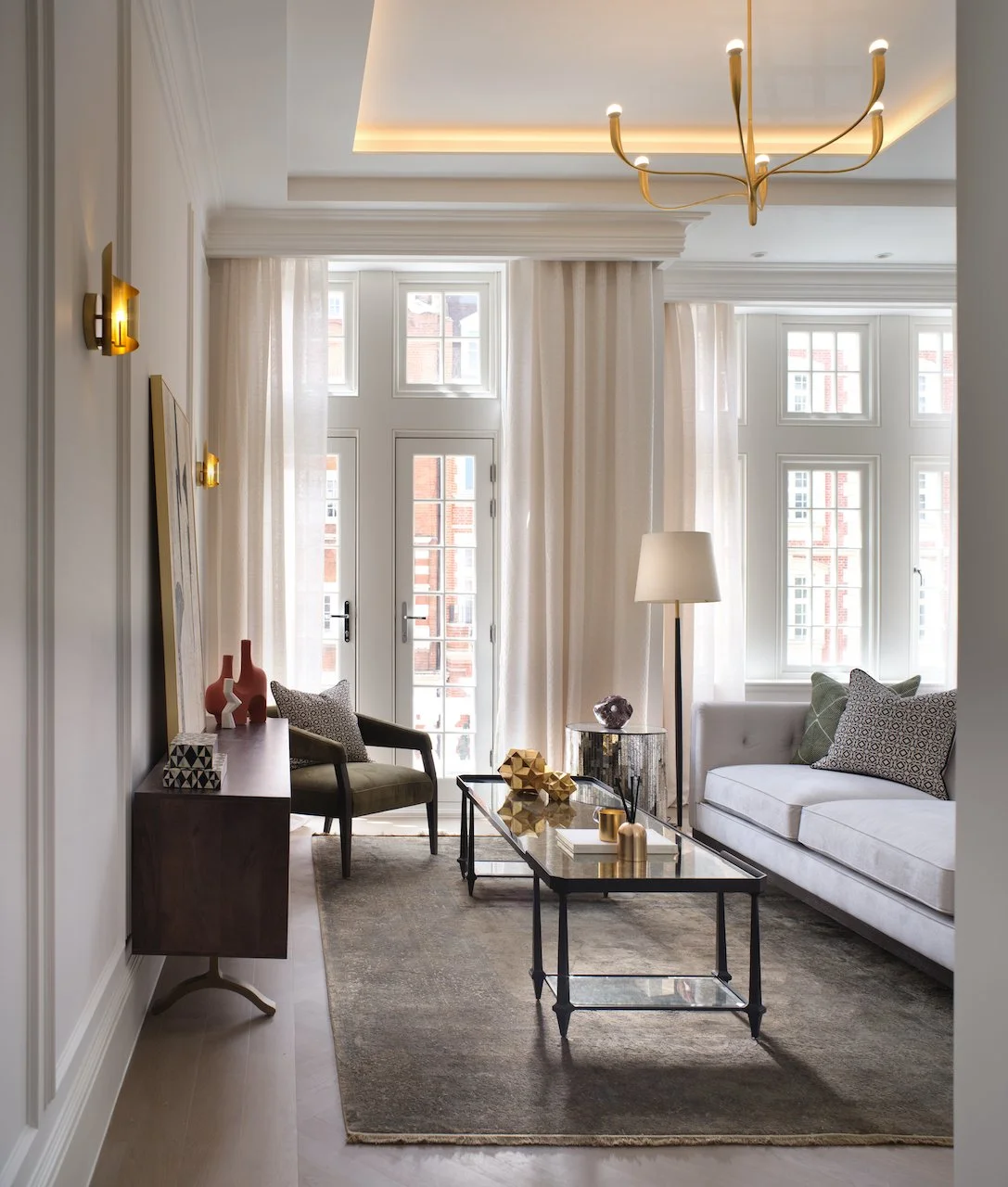 Elegant living room with large windows, white curtains, a white sofa with patterned pillows, a wooden armchair, a tall floor lamp, a glass coffee table with decorative objects, and a gold chandelier.