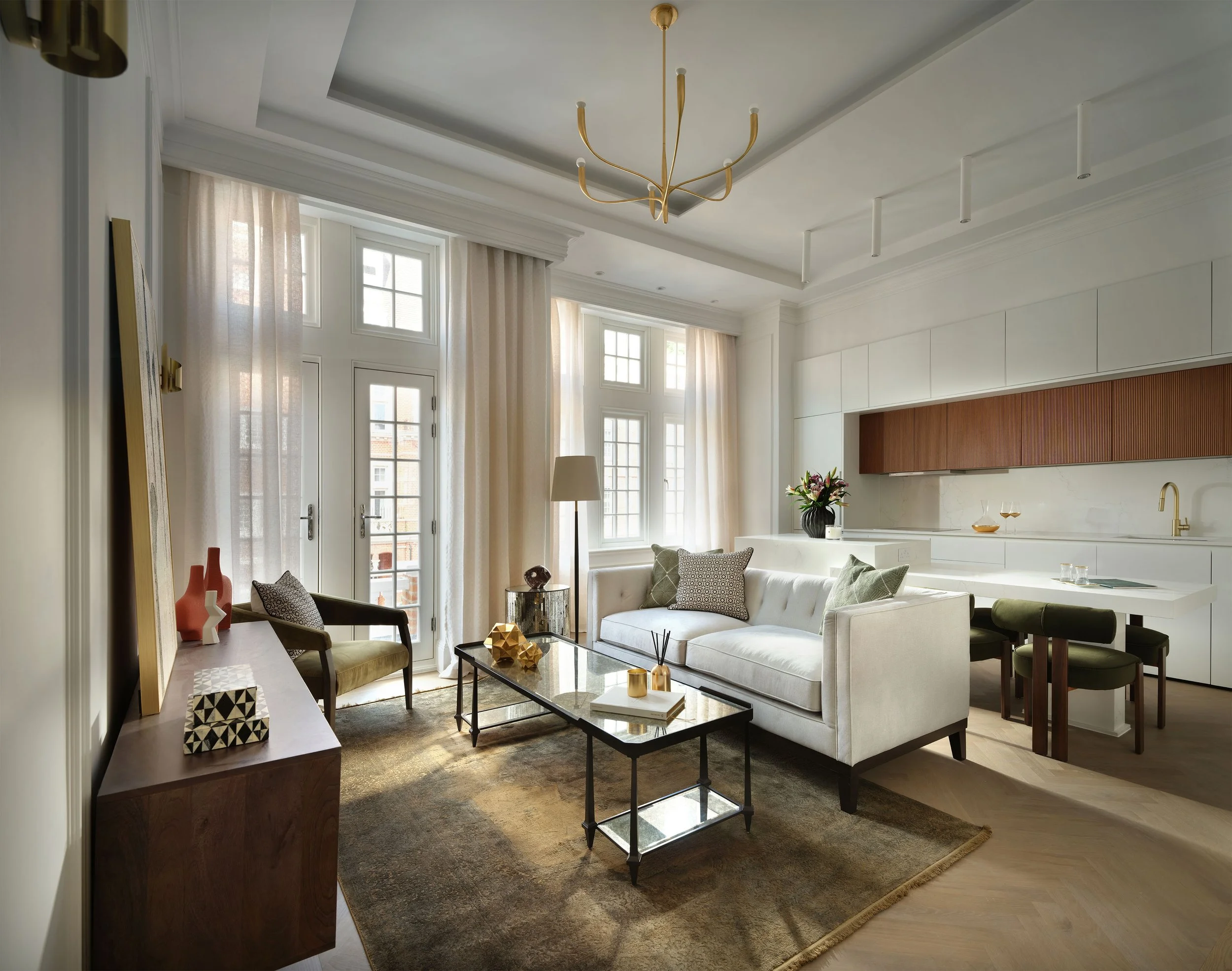 Living room with cream sofa, green chairs, glass coffee table, large windows with beige curtains, modern kitchen area with white cabinets and wooden accents