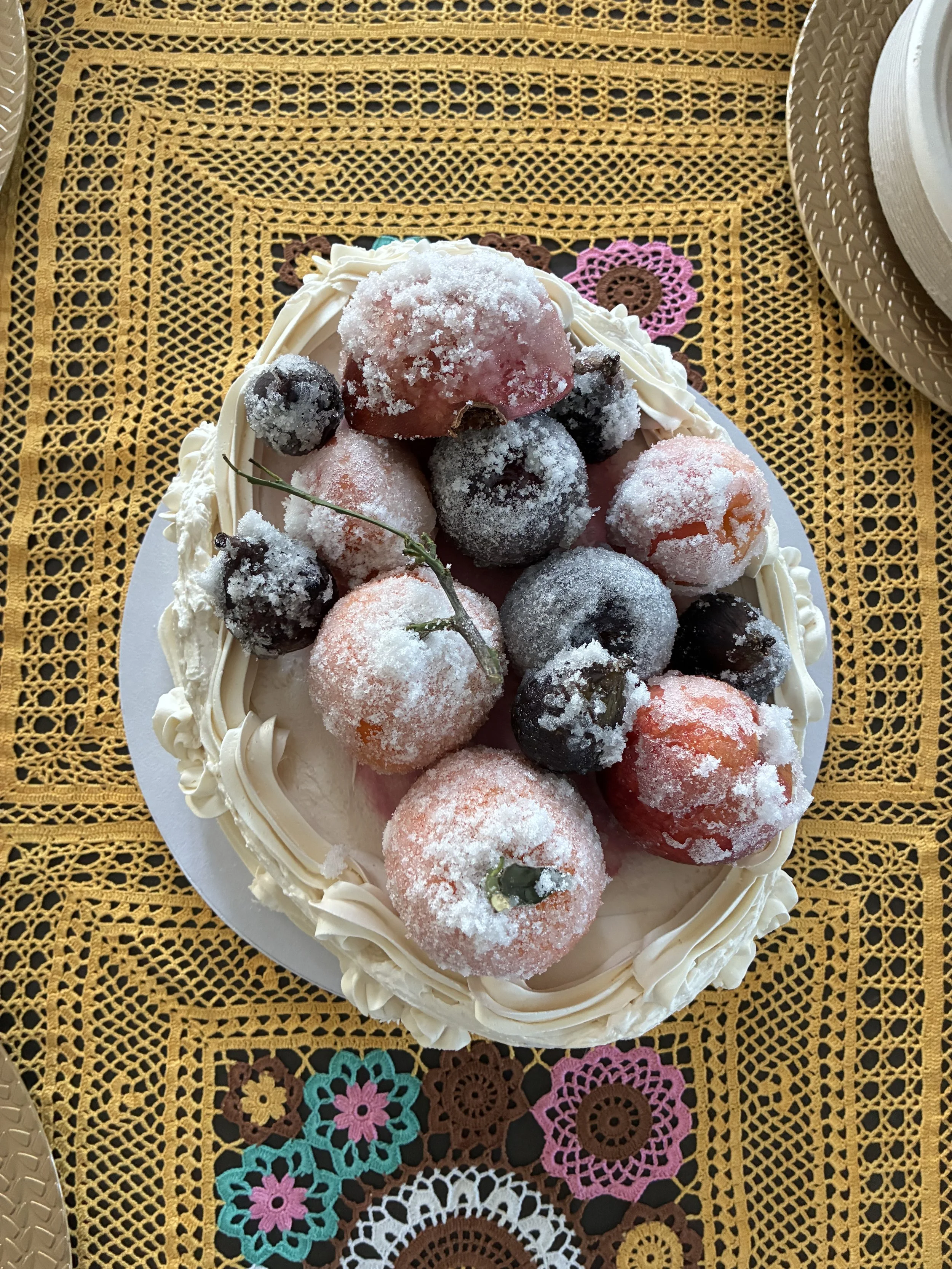 A cake decorated with strawberries and blueberries, dusted with powdered sugar, on a table with a yellow crochet tablecloth.
