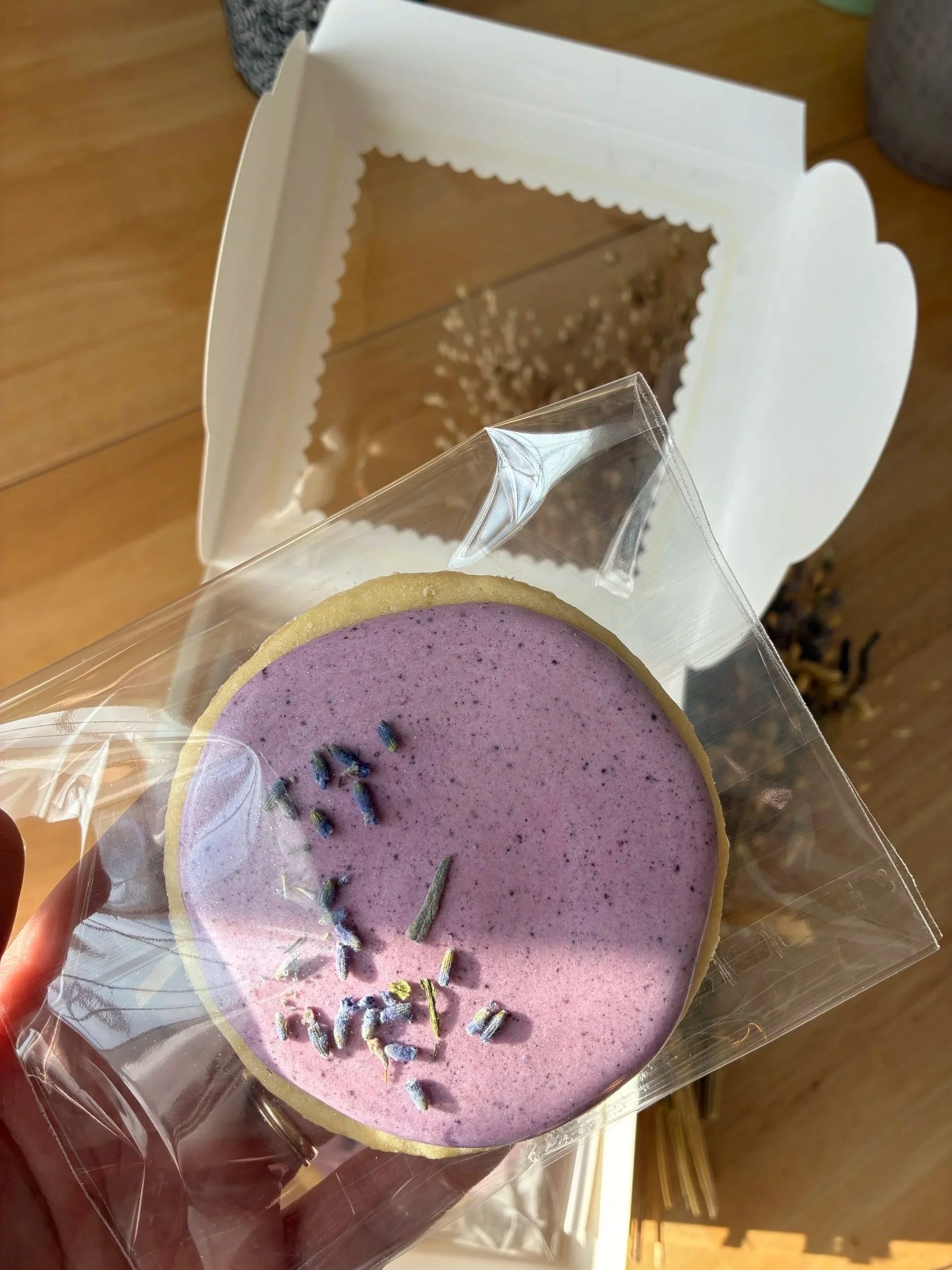 A hand holding a heart-shaped lavender-colored soap with sprigs of lavender on top, wrapped in clear plastic, in front of a white box with a mirror.