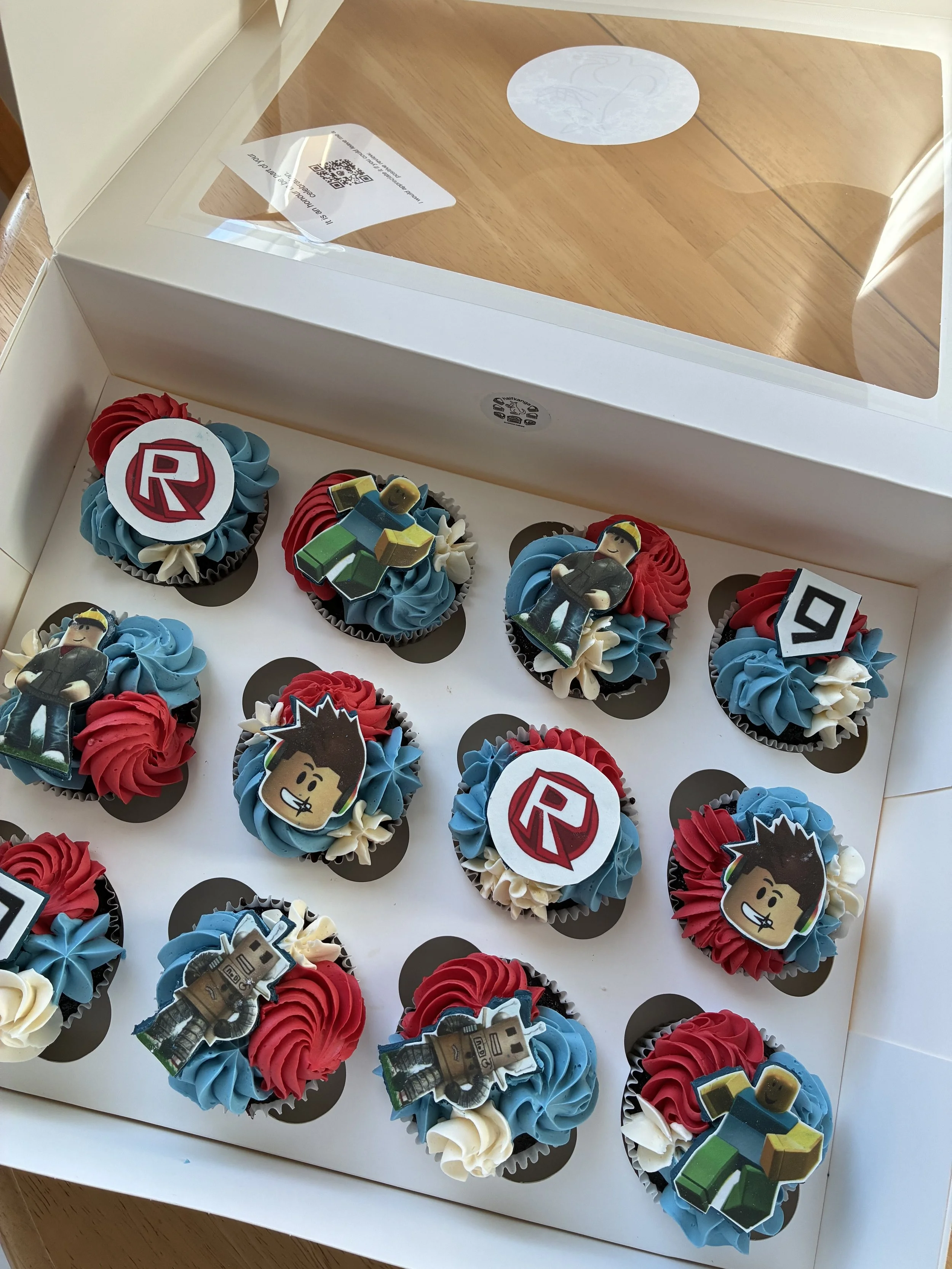 A box of decorated cupcakes with red, white, and blue swirl frosting and Roblox-themed toppers, including the Roblox logo, characters, and a robot, inside a white box with a clear lid.