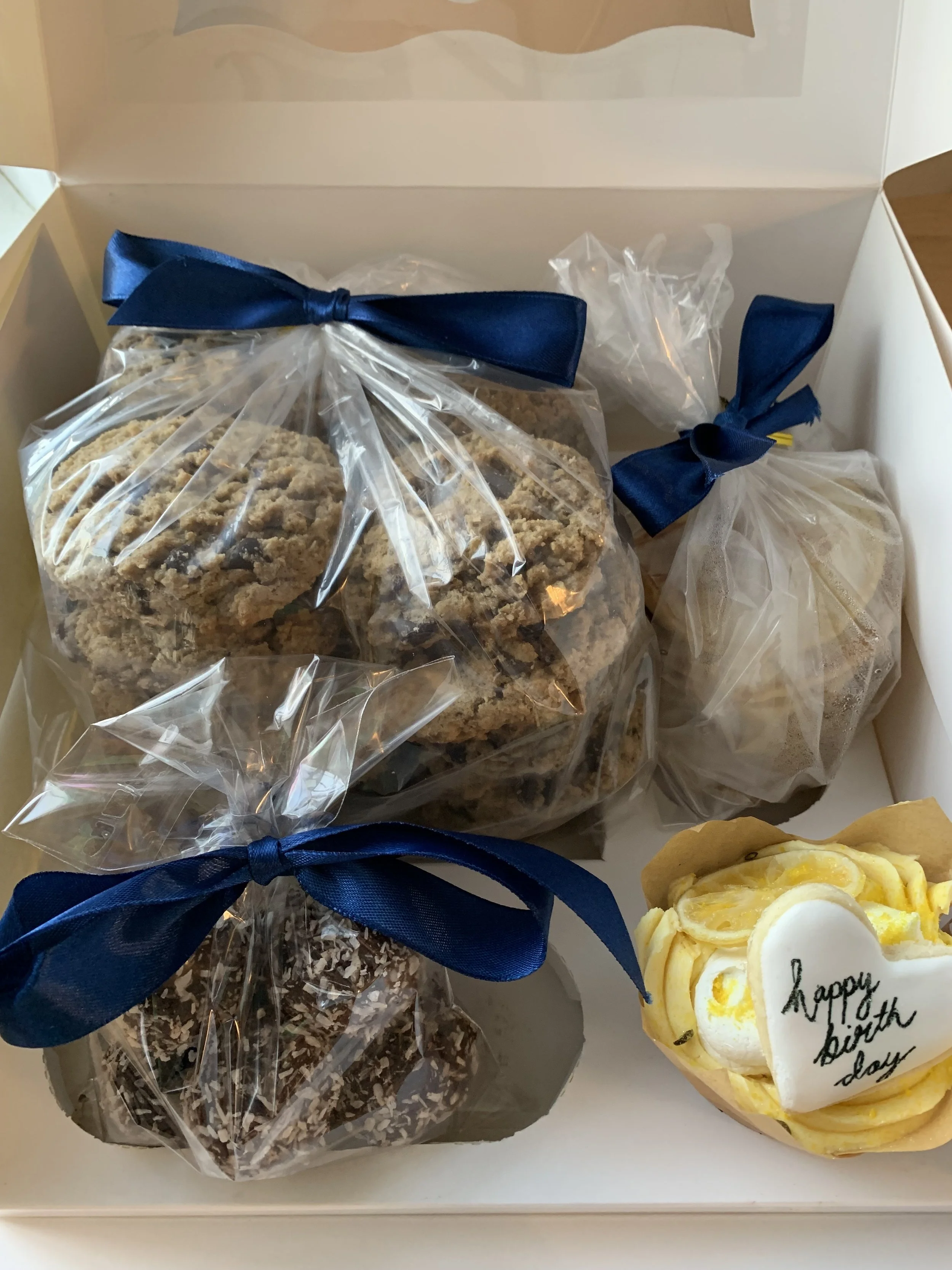 A gift box containing wrapped cookies, a chocolate treat, and a lemon cake with "happy birthday" written on a white chocolate plaque.