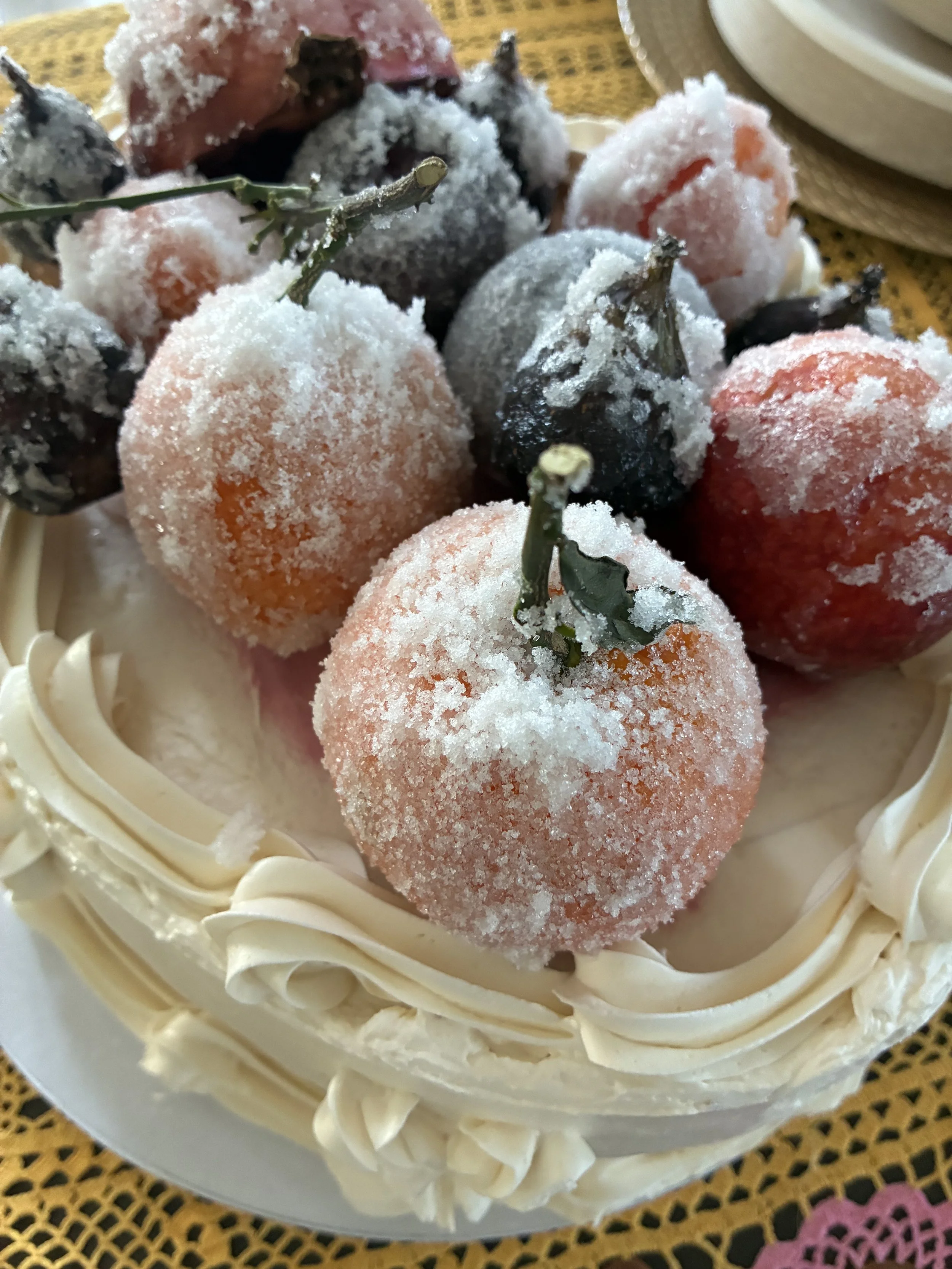 A cake decorated with frosted snowball-shaped fruits such as peaches, plums, and berries, with white cream frosting around the edges on a decorative table.