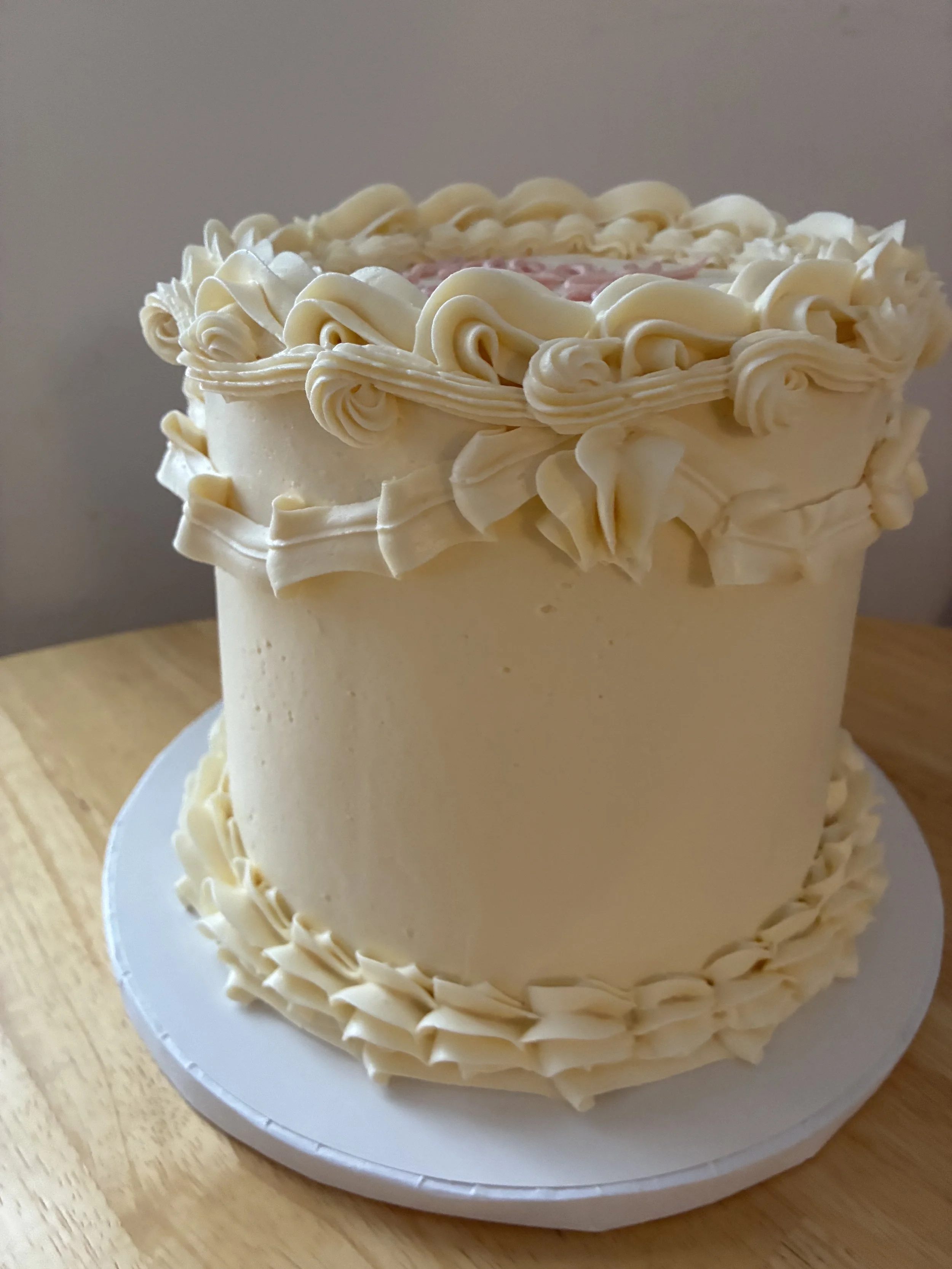 Cream-colored birthday cake decorated with piped frosting rosettes and borders, placed on a white cake board on a wooden surface.