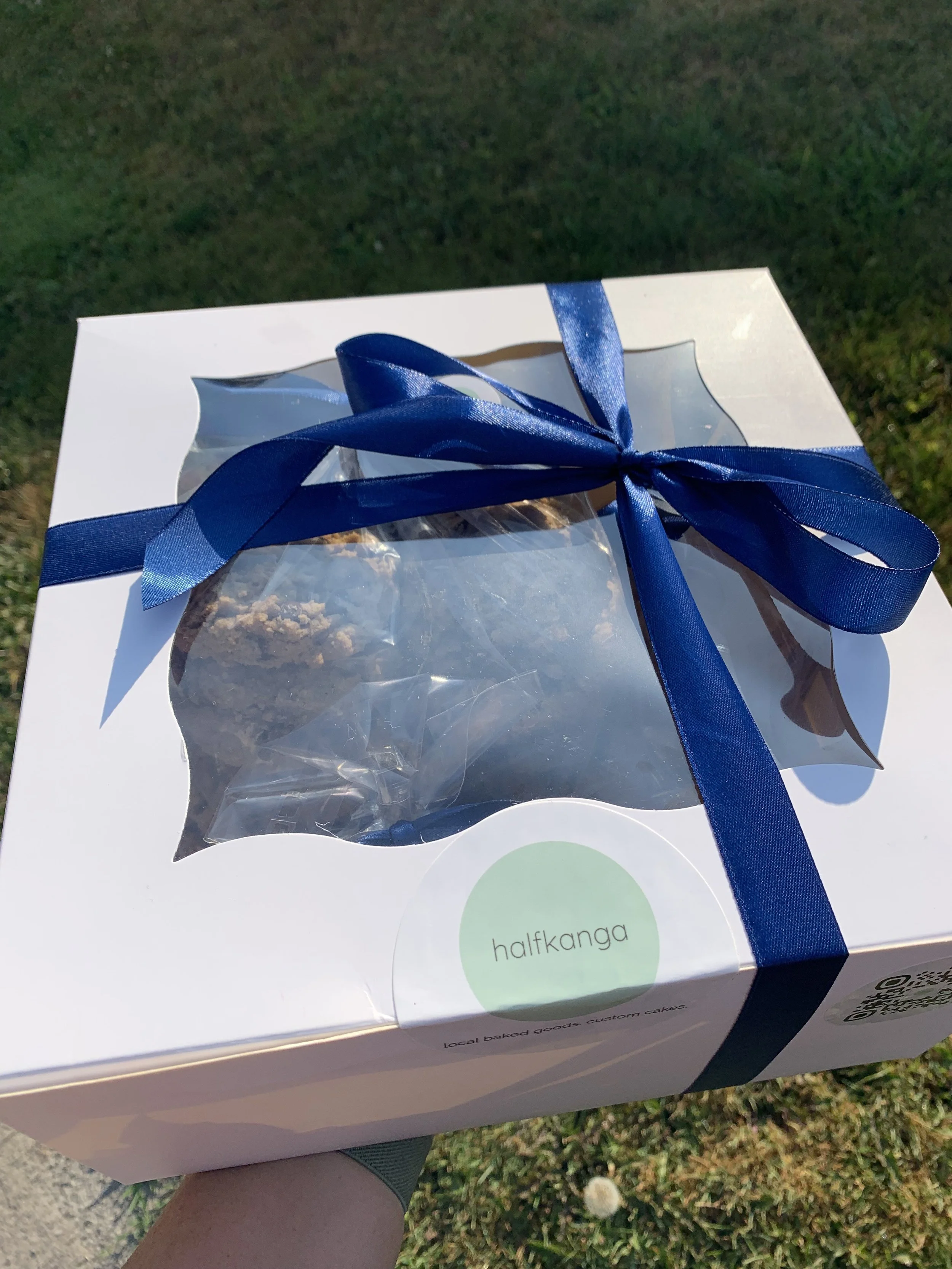A white box with a clear plastic window showing baked goods inside, tied with a blue ribbon, with a round label that says 'halfkanga', held outdoors over grass.