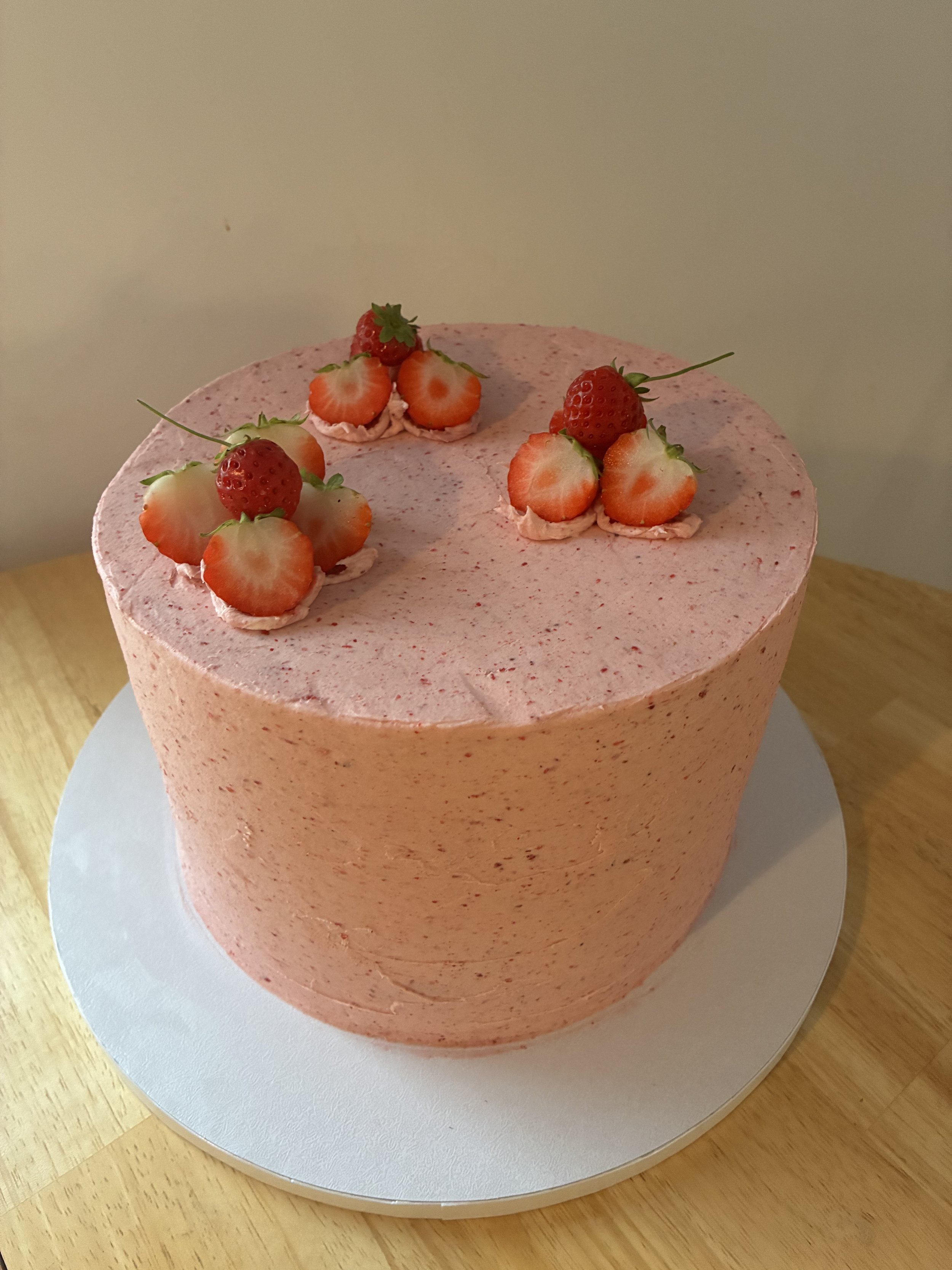 A pink frosted cake with strawberry decorations on top, placed on a silver cake board on a wooden surface.