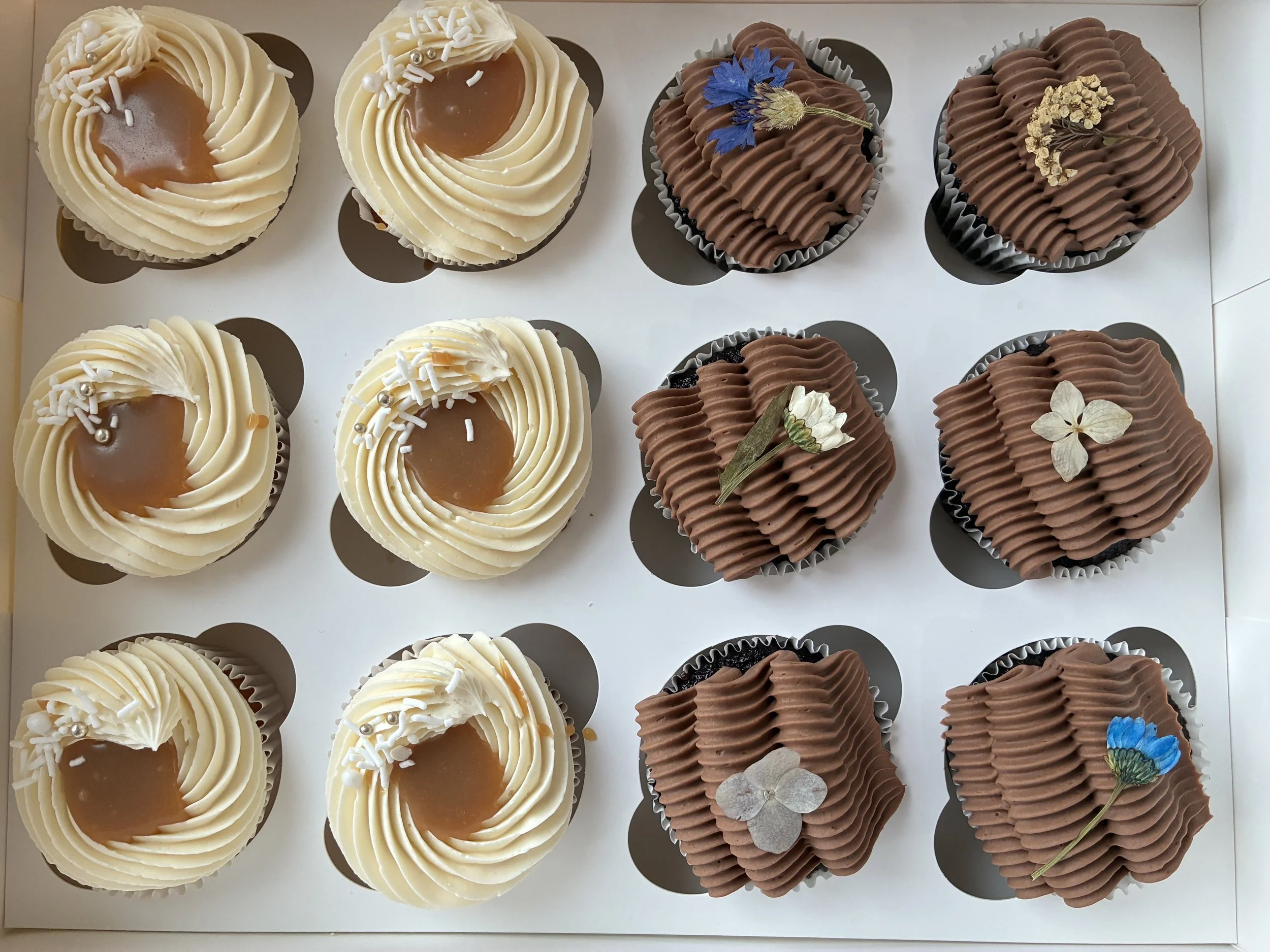 Box of twelve cupcakes, six with white frosting and caramel topping, six with chocolate frosting and edible flower decorations.