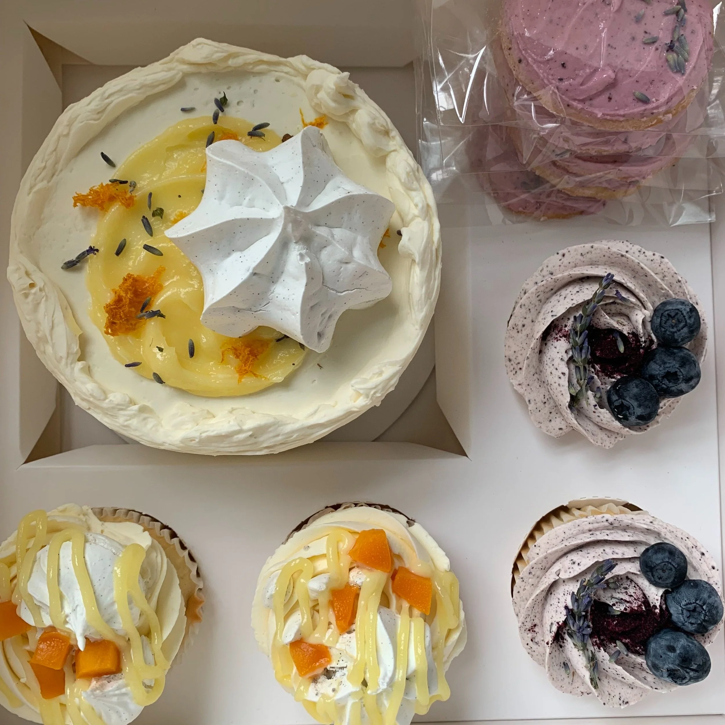A variety of decorated cupcakes and a cake, including a lemon-flavored cake with white frosting, a meringue topping, and garnishes like lavender and orange zest, two cupcakes with yellow drizzle, orange pieces, and white frosting, and two chocolate-flavored cupcakes with grayish frosting, blueberries, and lavender.