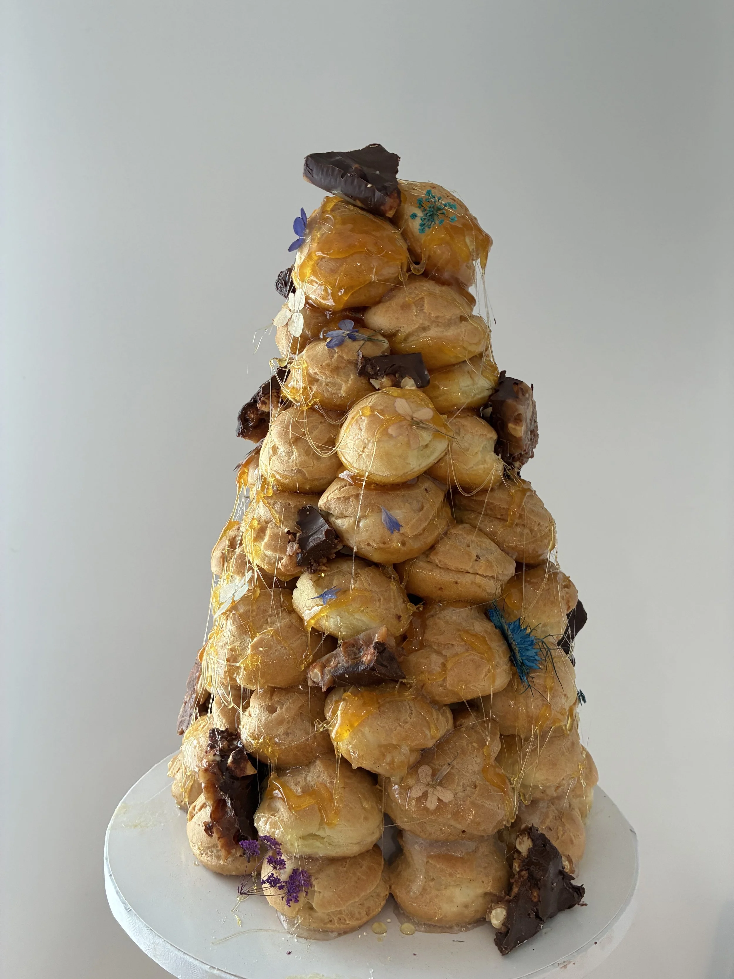 A pyramid-shaped dessert made of profiteroles topped with caramel, chocolate pieces, and edible flowers.