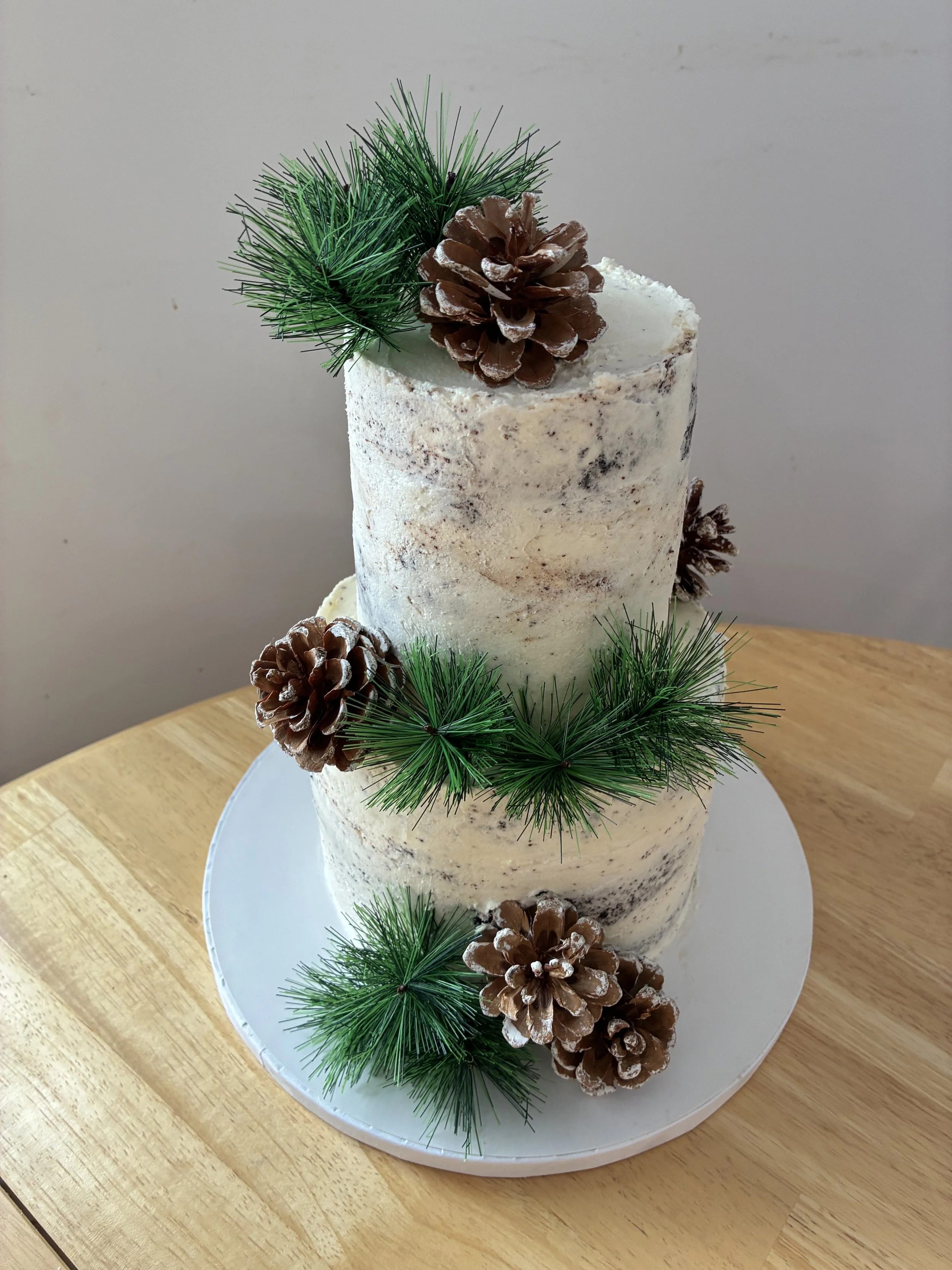 Two-layer white frosted cake decorated with pine cones and green pine branch edible decorations, placed on a white round cake board on a wooden table.