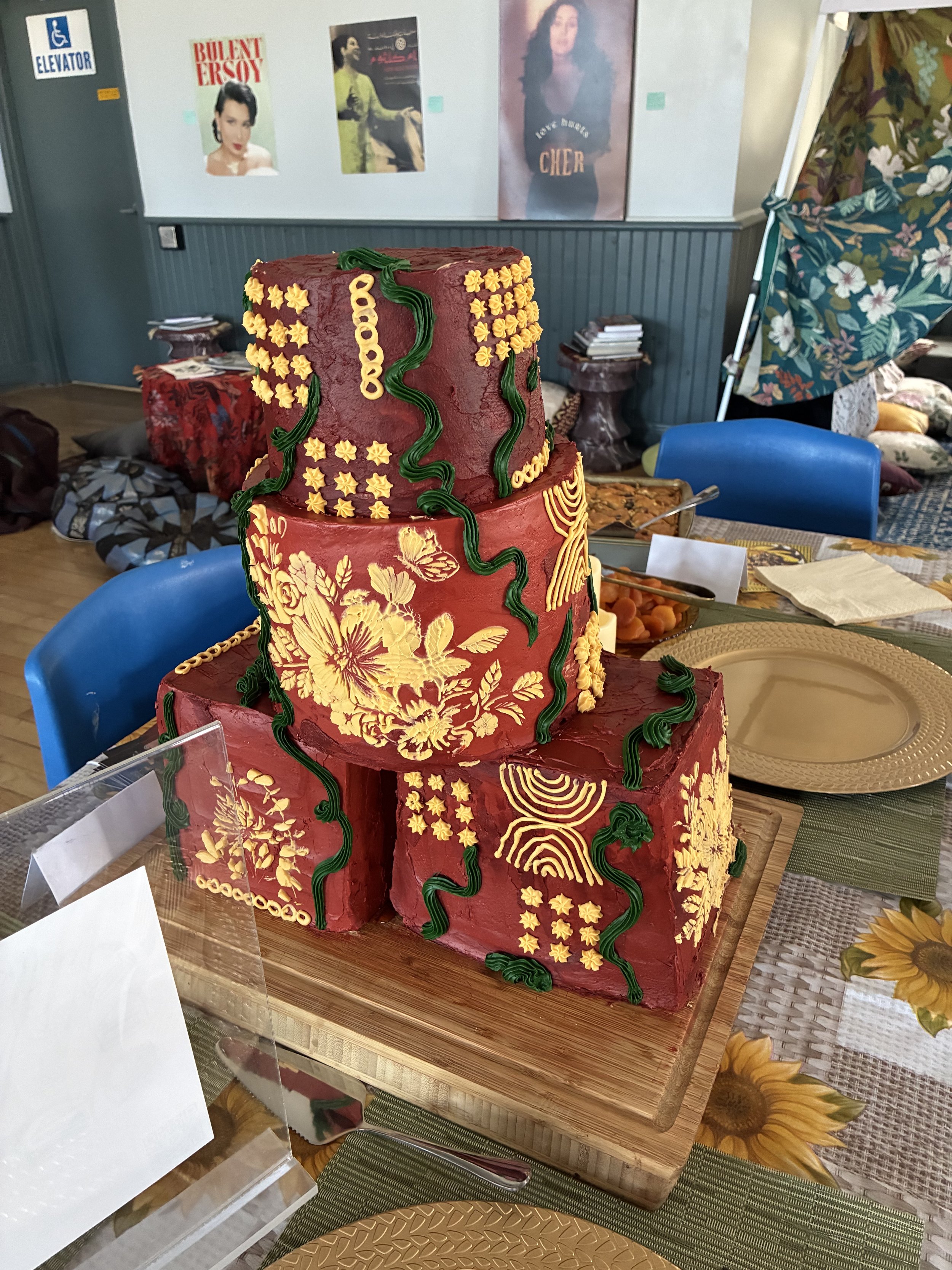 A three-tiered cake with a red base color, decorated with yellow floral and geometric patterns, and green wavy icing accents, placed on a wooden board on a table with sunflower-themed tablecloth.
