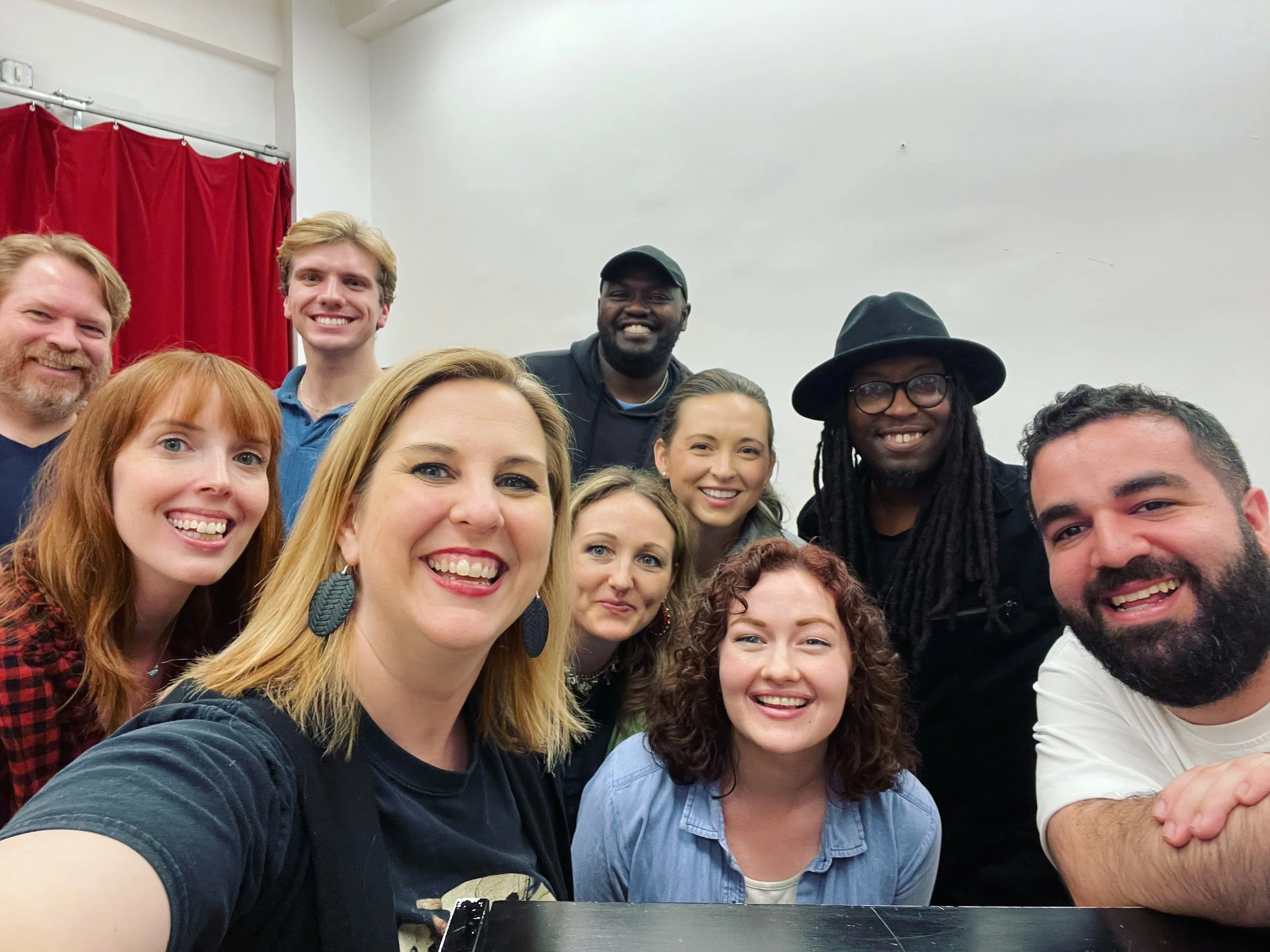 Group of ten smiling people taking a selfie in a room with white walls and a red curtain.