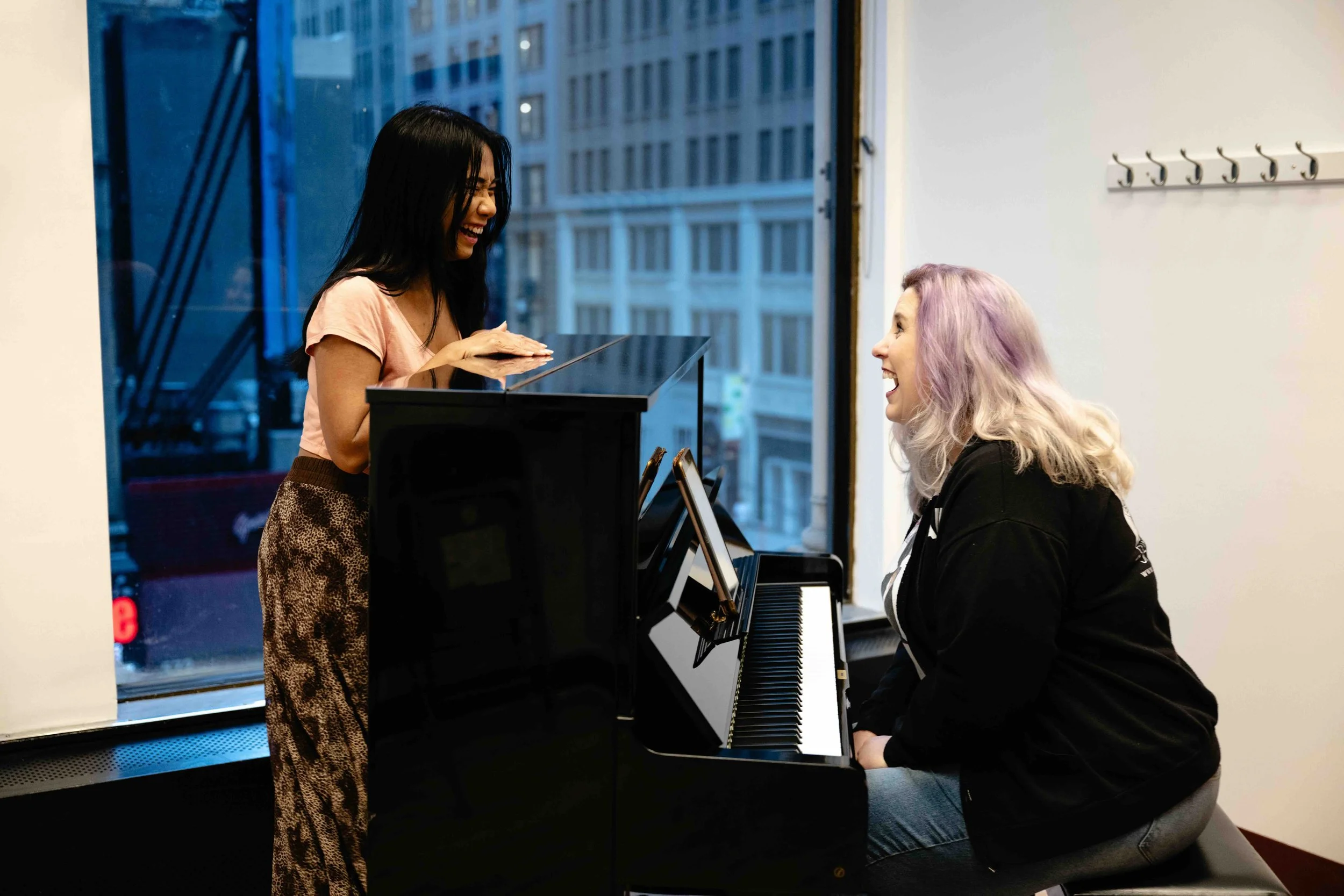 Two women laughing and enjoying a singing lesson in a bright room with large windows, one standing and the other sitting at the piano.
