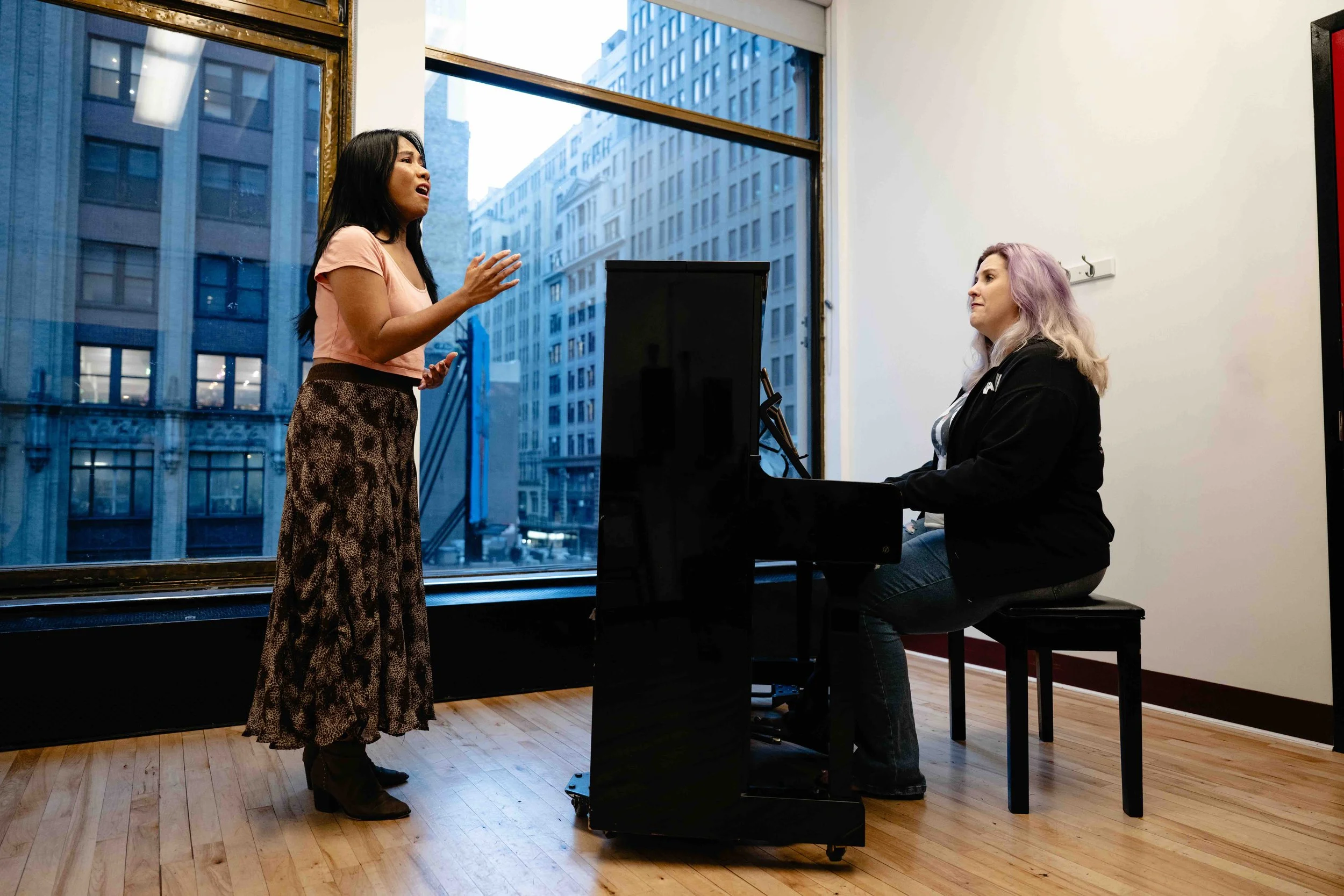 A woman with long dark hair, wearing a pink shirt and patterned pants, is singing passionately while standing in front of a large window. Another woman with shoulder-length blonde hair, wearing a black jacket, is sitting at a black piano, listening attentively inside a room with wooden flooring and city buildings visible outside through the window.